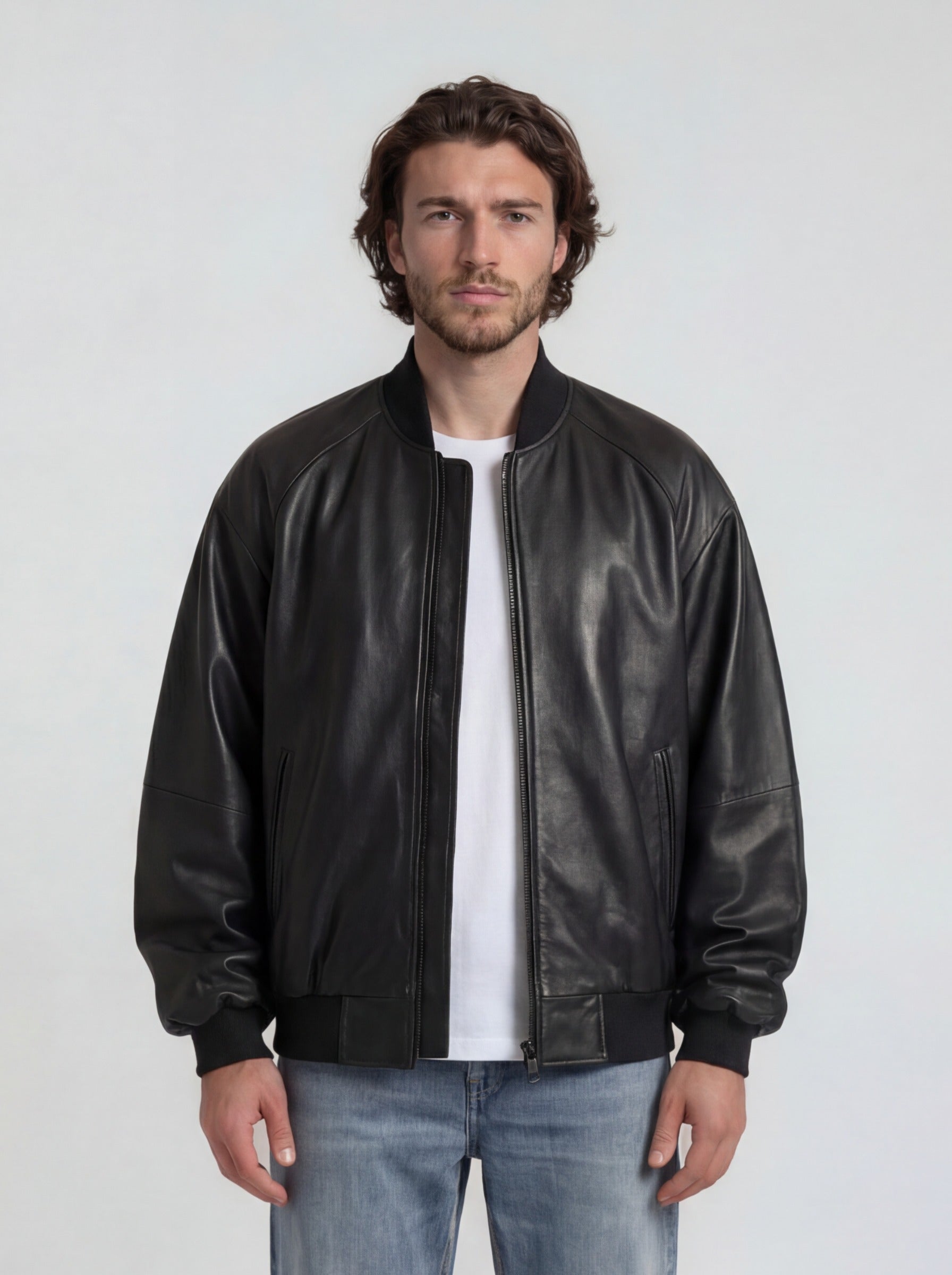 Ein Mann mit gewellten braunen Haaren und kurzem Bart steht vor einem schlichten Hintergrund und trägt die trueprodigy Bomber Lederjacke Herren "Filip" im Oversize-Look aus Schafsleder, gepaart mit einem weißen T-Shirt und hellblauen Jeans.