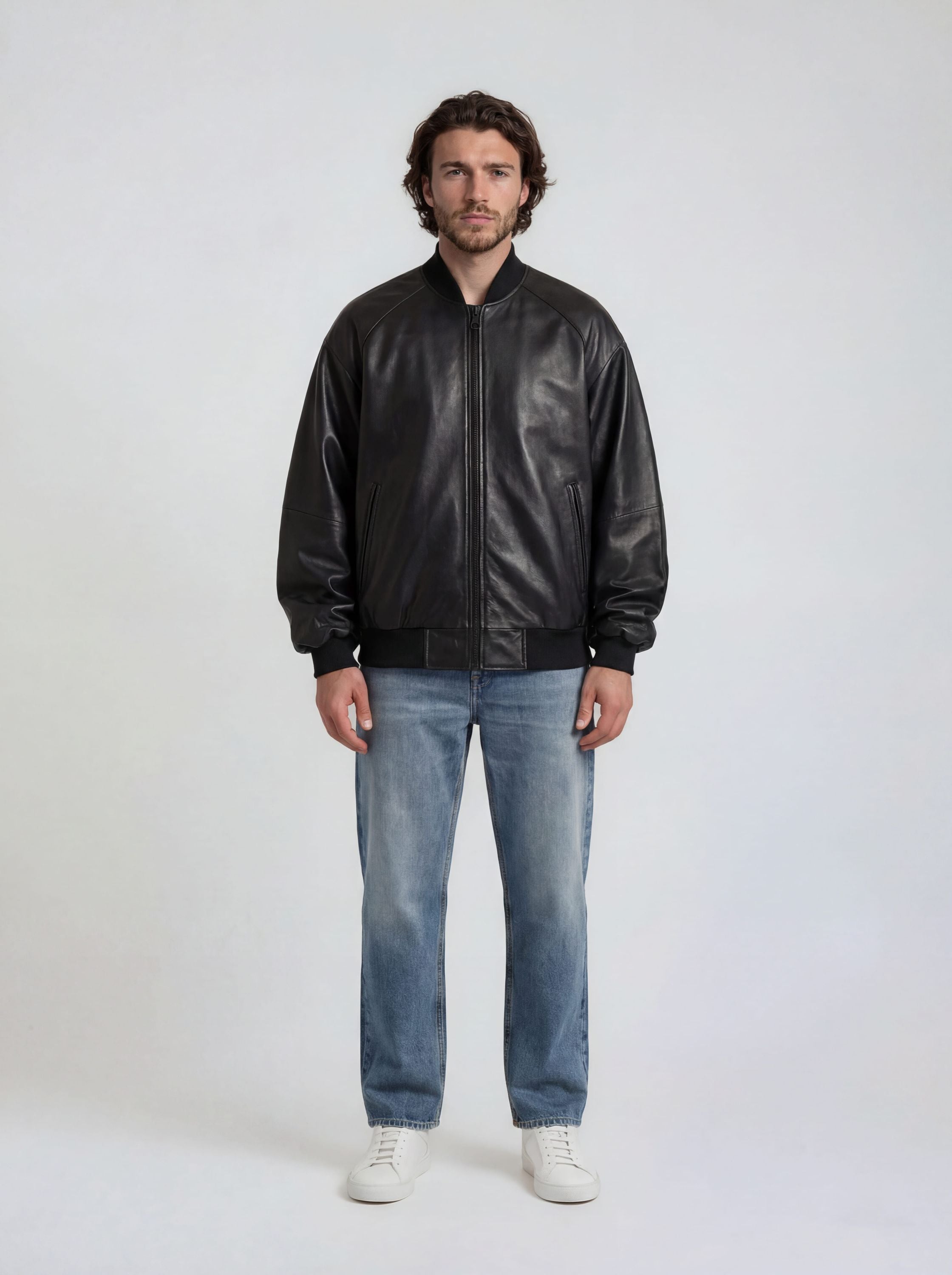 Ein Mann steht vor einem schlichten, hellen Hintergrund. Er trägt die trueprodigy Bomber Lederjacke Herren "Filip" im Oversize-Look, blaue Jeans und weiße Sneakers.