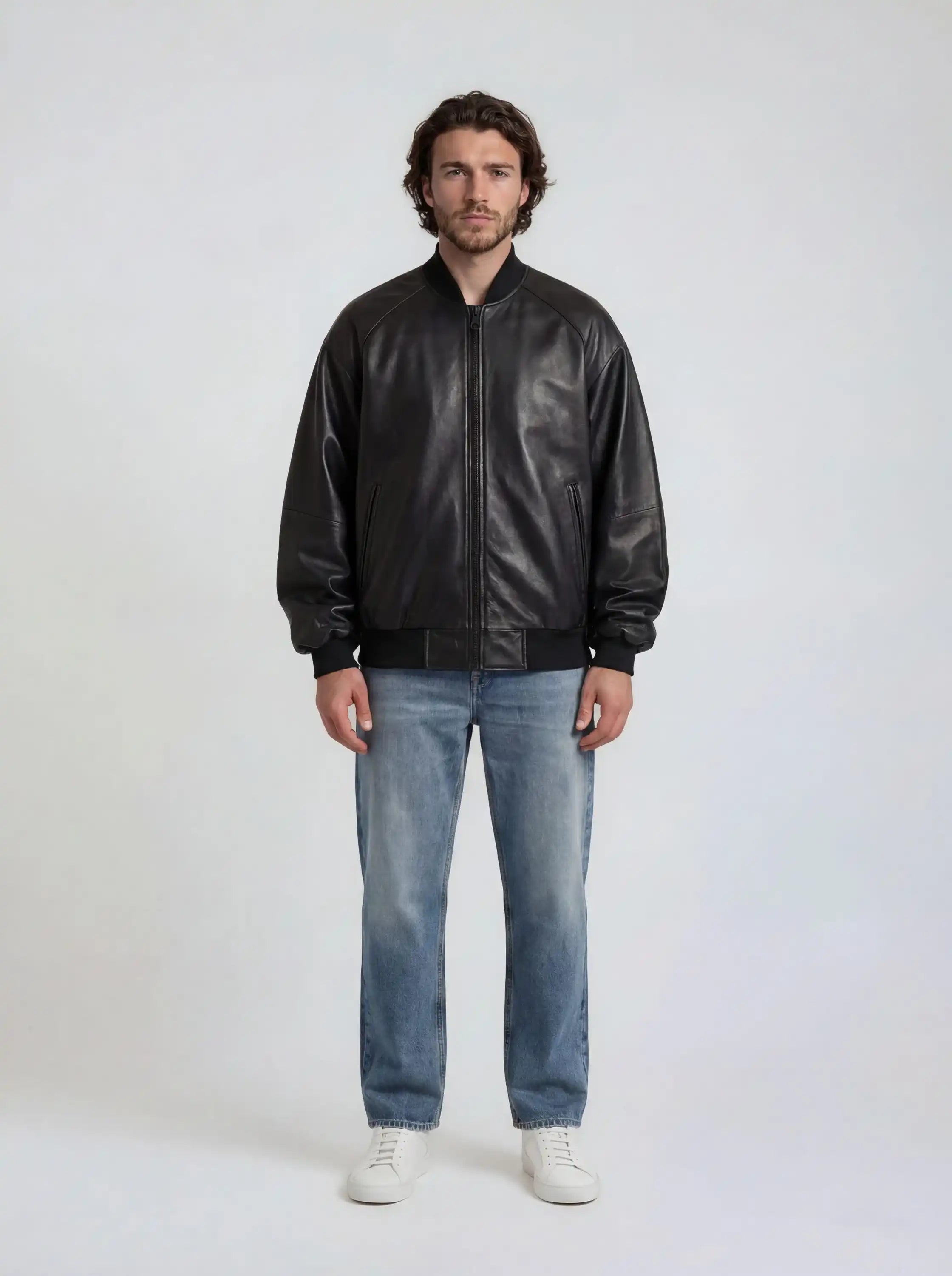 Ein Mann steht vor einem schlichten, hellen Hintergrund. Er trägt die trueprodigy Bomber Lederjacke Herren "Filip" im Oversize-Look, blaue Jeans und weiße Sneakers.