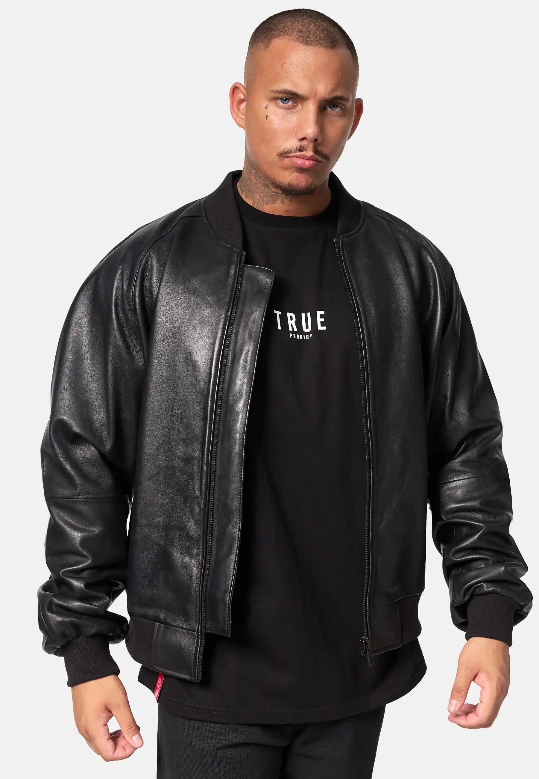 Ein Mann mit kurzen Haaren und gestutztem Bart trägt eine schwarze trueprodigy Filip Oversized Bomber-Lederjacke über einem schwarzen Hemd mit dem Aufdruck "TRUE", stehend vor einem schlichten hellen Hintergrund.
