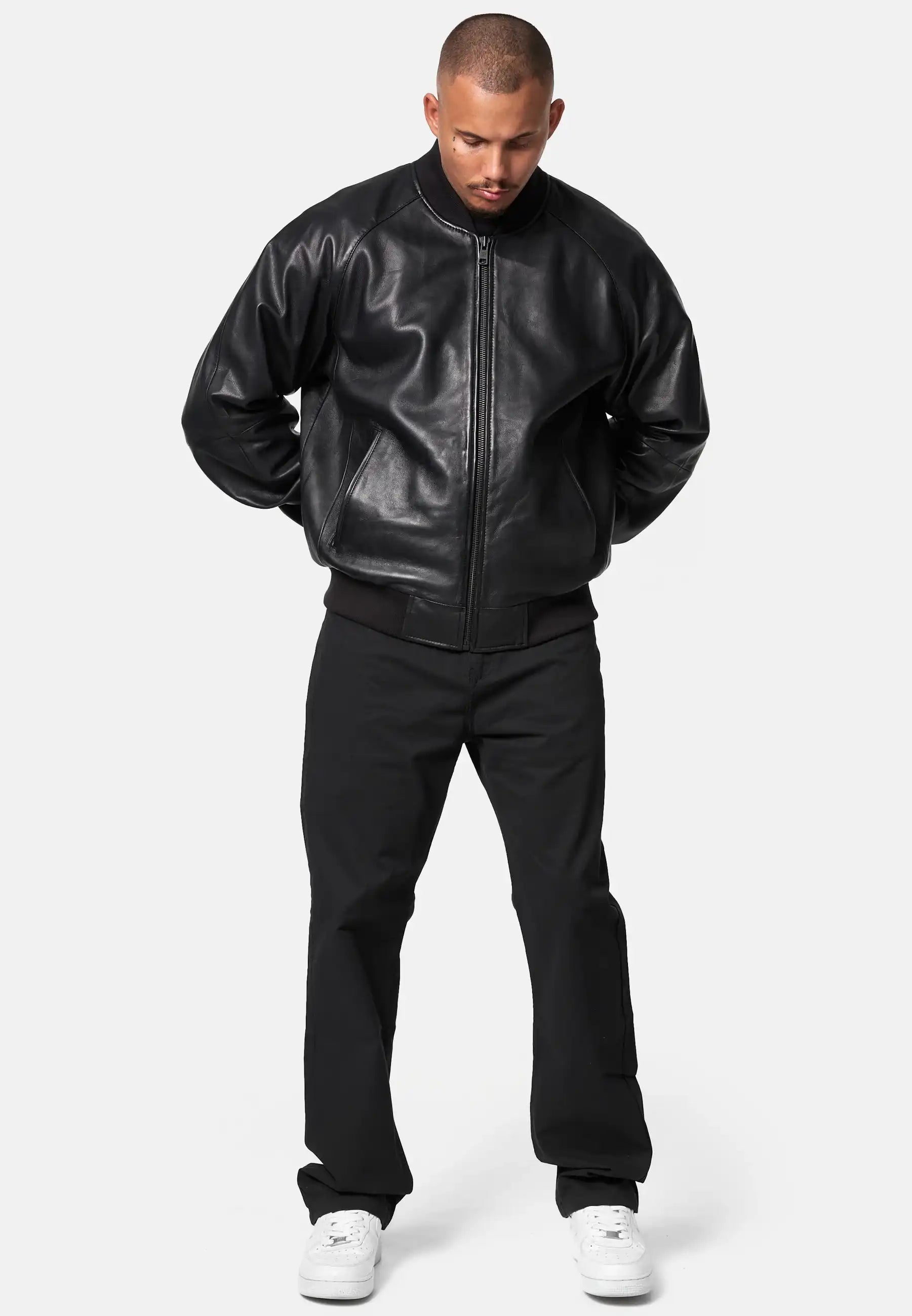 Ein Mann blickt mit leicht gesenktem Kopf nach vorne, die Hände auf dem Rücken, und trägt die trueprodigy Filip Oversized Bomber-Lederjacke aus schwarzem Leder, eine schwarze Hose und weiße Turnschuhe vor einem schlichten hellen Hintergrund.