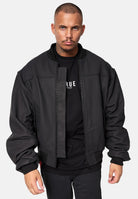 Ein Mann mit Kurzhaarschnitt und Schnurrbart trägt eine schwarze Nazar Oversized Bomber-Lederjacke matt von trueprodigy über einem schwarzen trueprodigy-Shirt mit weißem Text, steht vor einem schlichten weißen Hintergrund und blickt mit neutralem Ausdruck nach vorne.