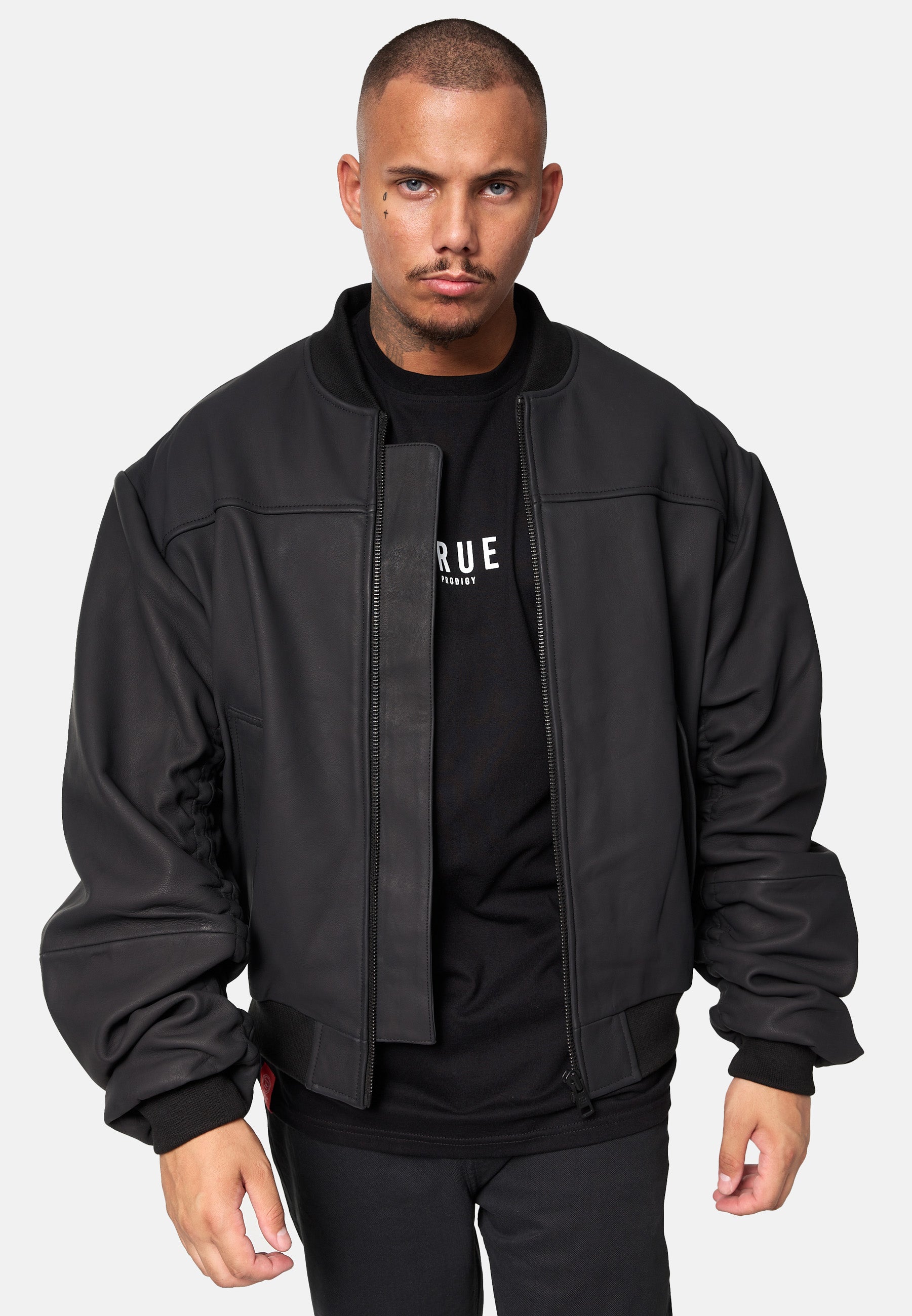 Ein Mann mit Kurzhaarschnitt und Schnurrbart trägt eine schwarze Nazar Oversized Bomber-Lederjacke matt von trueprodigy über einem schwarzen trueprodigy-Shirt mit weißem Text, steht vor einem schlichten weißen Hintergrund und blickt mit neutralem Ausdruck nach vorne.