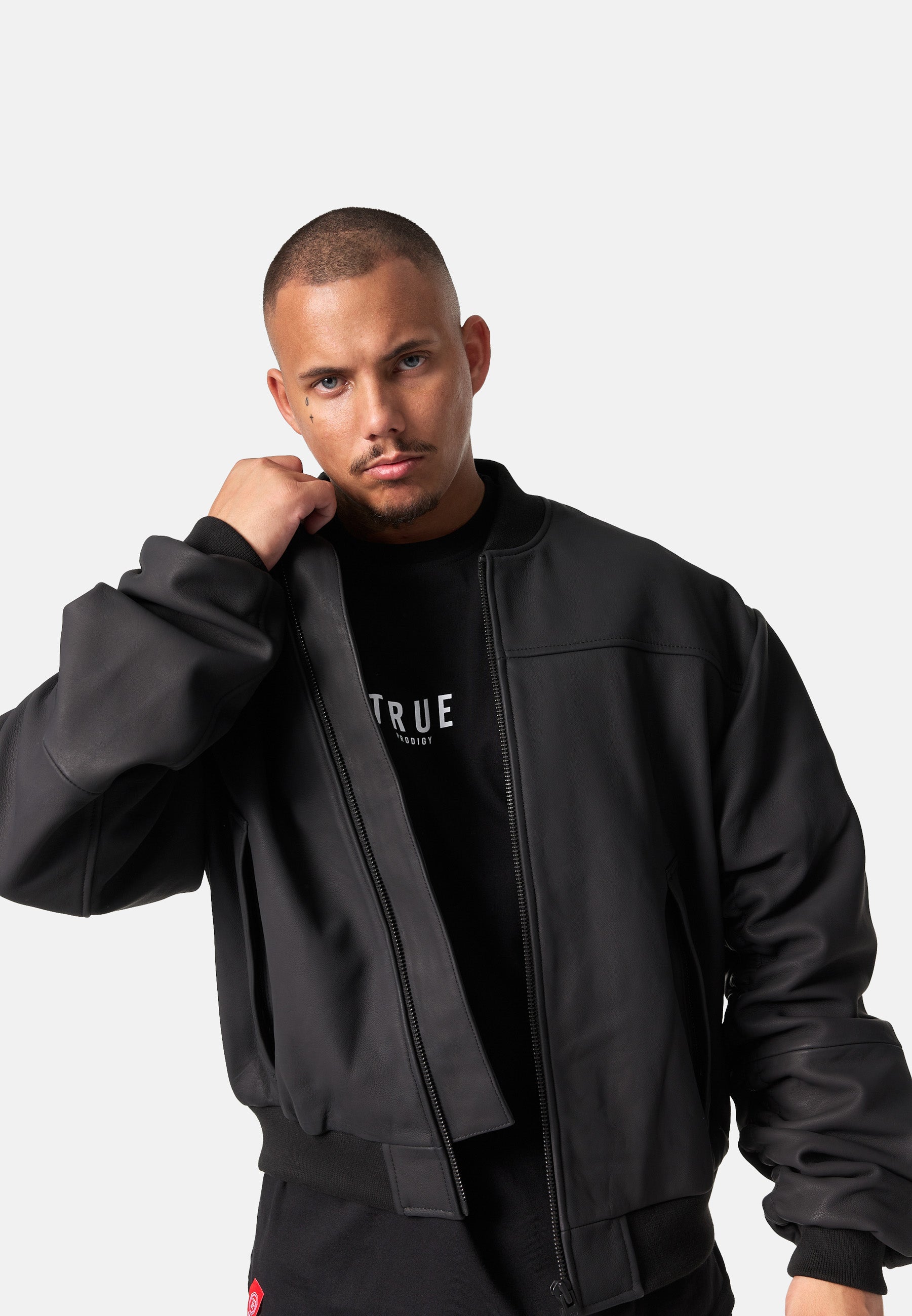 Ein Mann mit kurzem Haar und getrimmtem Bart trägt die Nazar Oversized Bomber-Lederjacke matt von trueprodigy offen über einem schwarzen Shirt mit weißer Schrift und blickt direkt in die Kamera. Der Hintergrund ist schlicht weiß.