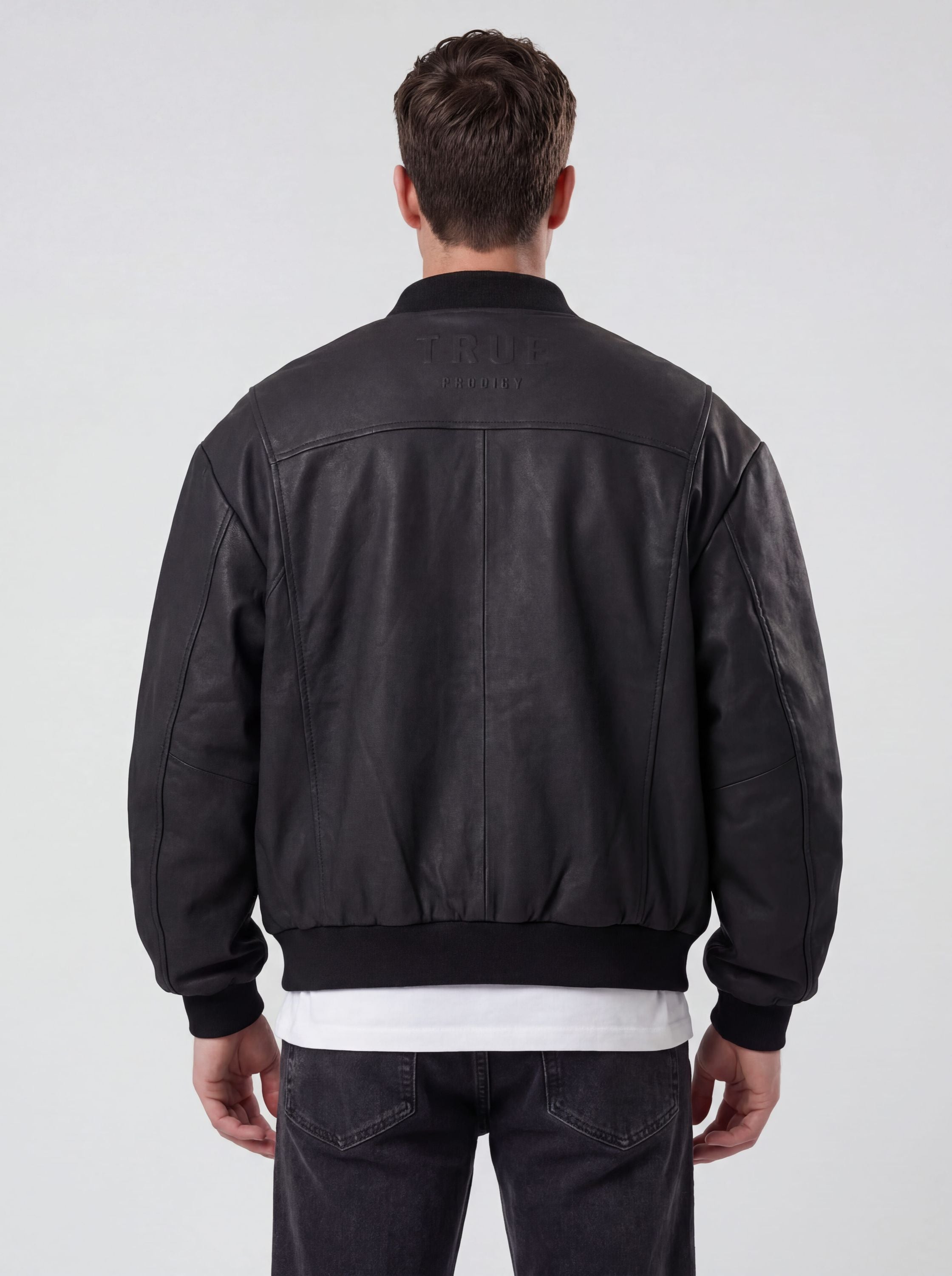 Ein Mann steht mit dem Rücken zur Kamera und trägt die trueprodigy Bomber Lederjacke Herren "Nazar" im Oversize-Schnitt in schwarz, über einem weißen Hemd und einer dunklen Jeans. Der Hintergrund ist einfarbig und hell gefärbt.