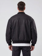 Ein Mann steht mit dem Rücken zur Kamera und trägt die trueprodigy Bomber Lederjacke Herren "Nazar" im Oversize-Schnitt in schwarz, über einem weißen Hemd und einer dunklen Jeans. Der Hintergrund ist einfarbig und hell gefärbt.