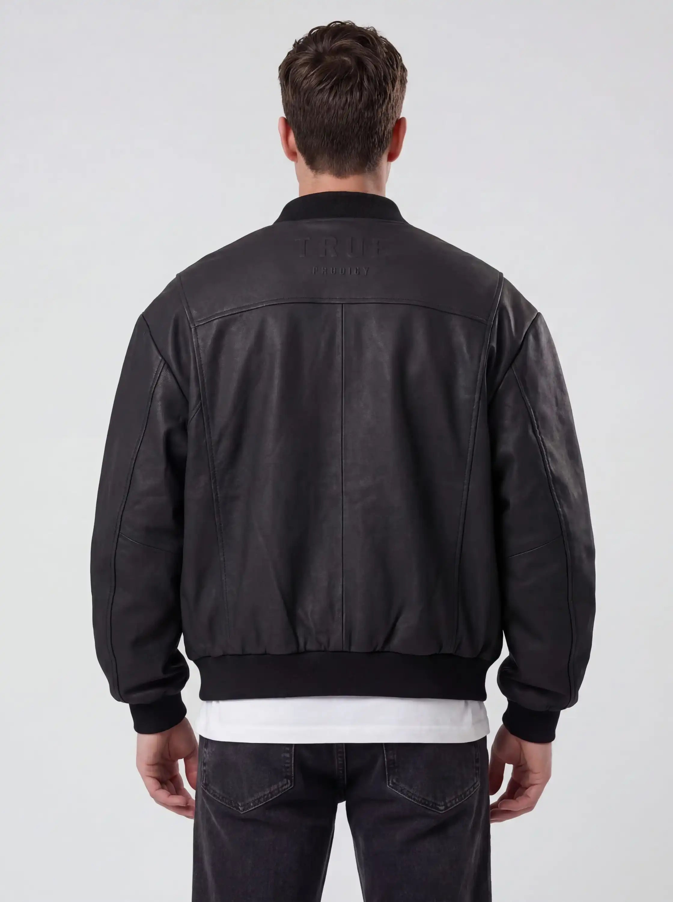 Ein Mann steht mit dem Rücken zur Kamera und trägt die trueprodigy Bomber Lederjacke Herren "Nazar" im Oversize-Schnitt in schwarz, über einem weißen Hemd und einer dunklen Jeans. Der Hintergrund ist einfarbig und hell gefärbt.
