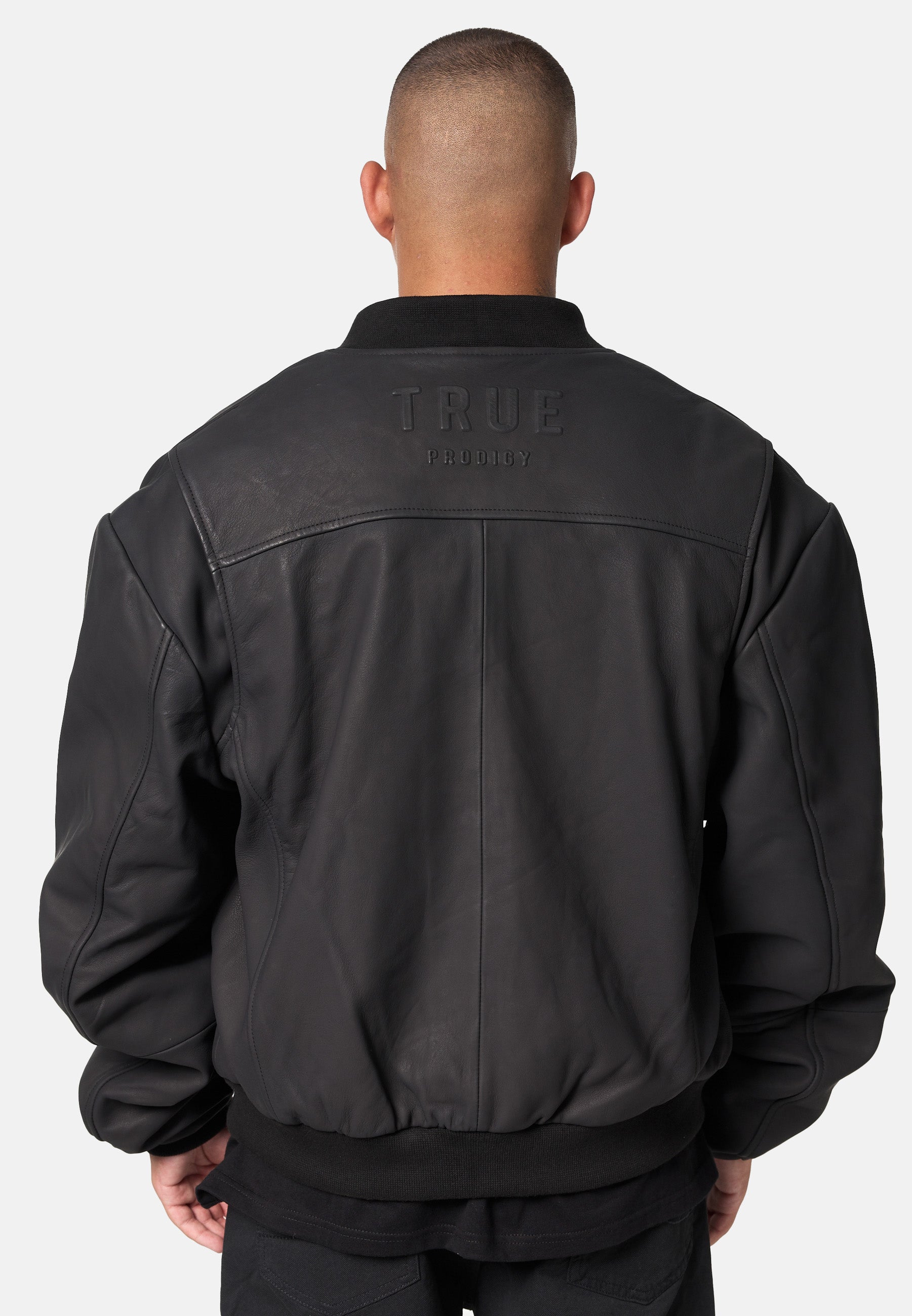 Eine Person mit kurzen Haaren, von hinten gesehen, trägt eine schwarze Nazar Oversized Bomber-Lederjacke matt von trueprodigy. Auf dem oberen Rücken ist "trueprodigy" vor einem schlichten hellen Hintergrund eingeprägt.