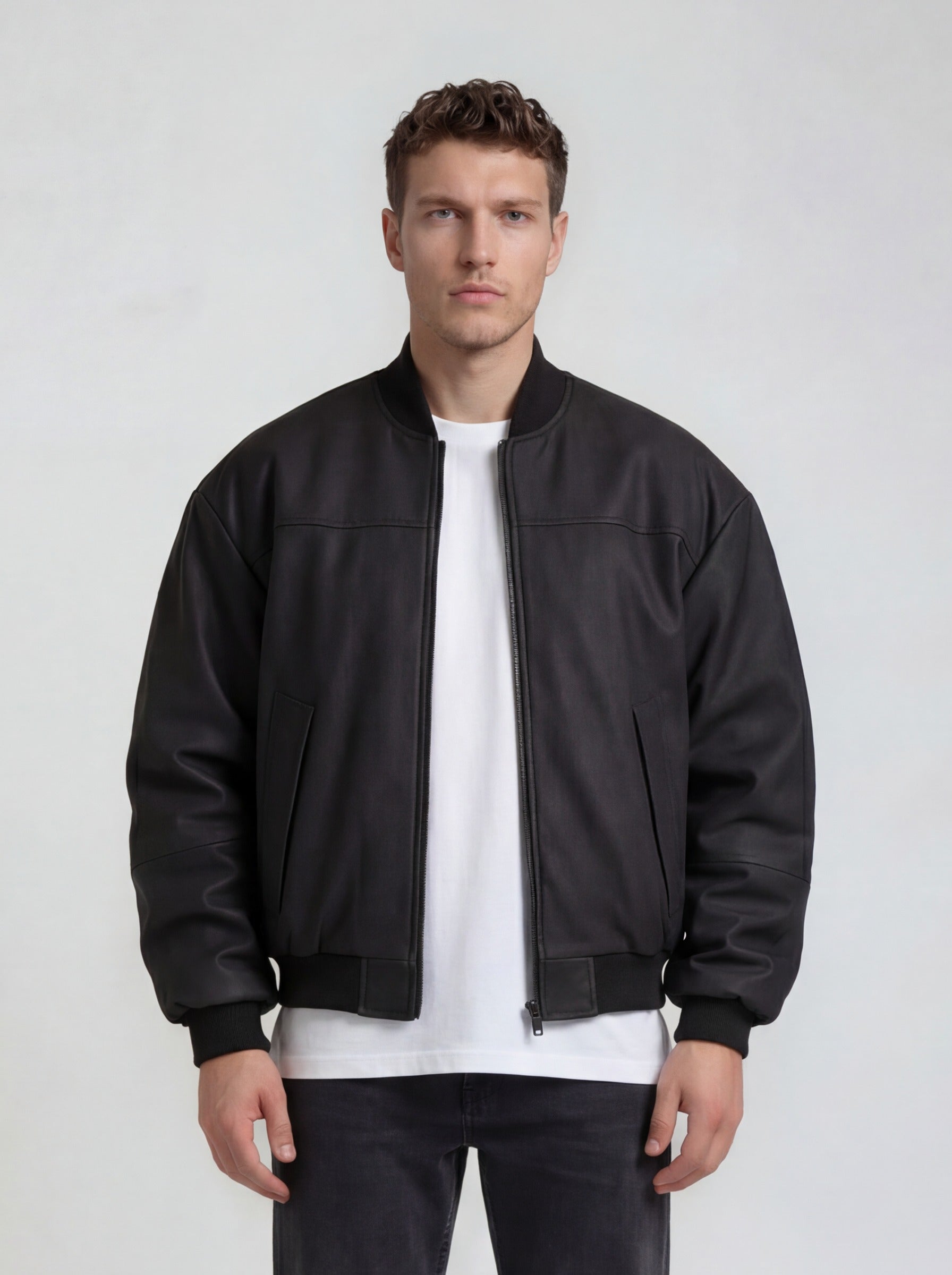 Ein Mann sieht nach vorne und trägt die trueprodigy Bomber Lederjacke Herren "Nazar" im Oversize-Schnitt in Schwarz über einem weißen T-Shirt und schwarzen Jeans vor hellem Hintergrund.