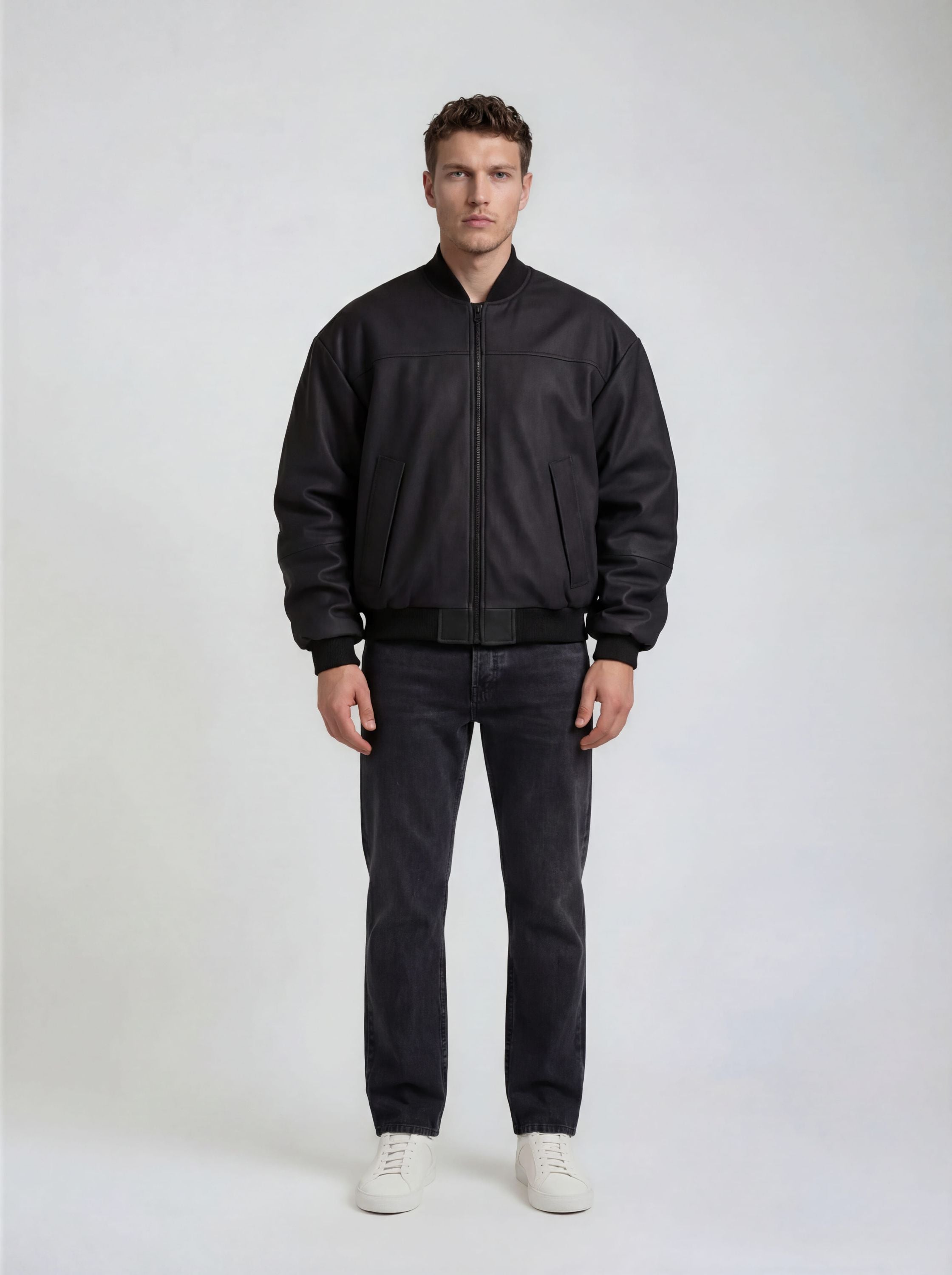 Ein Mann steht vor einem schlichten, hellen Hintergrund und trägt die trueprodigy Bomber Lederjacke Herren "Nazar" im Oversize-Schnitt, gepaart mit einer dunklen Jeans und weißen Sneakers. Seine Arme hängen entspannt an seinen Seiten und sein Gesichtsausdruck ist neutral.