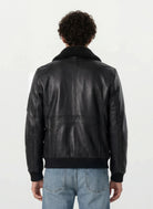 Eine Person mit kurzen lockigen Haaren ist von hinten zu sehen. Sie trägt eine Trueprodigy Moderne Fliegerjacke aus echtem Leder mit abnehmbarem Teddy-Kragen und hellblaue Jeans vor einem schlichten hellen Hintergrund.