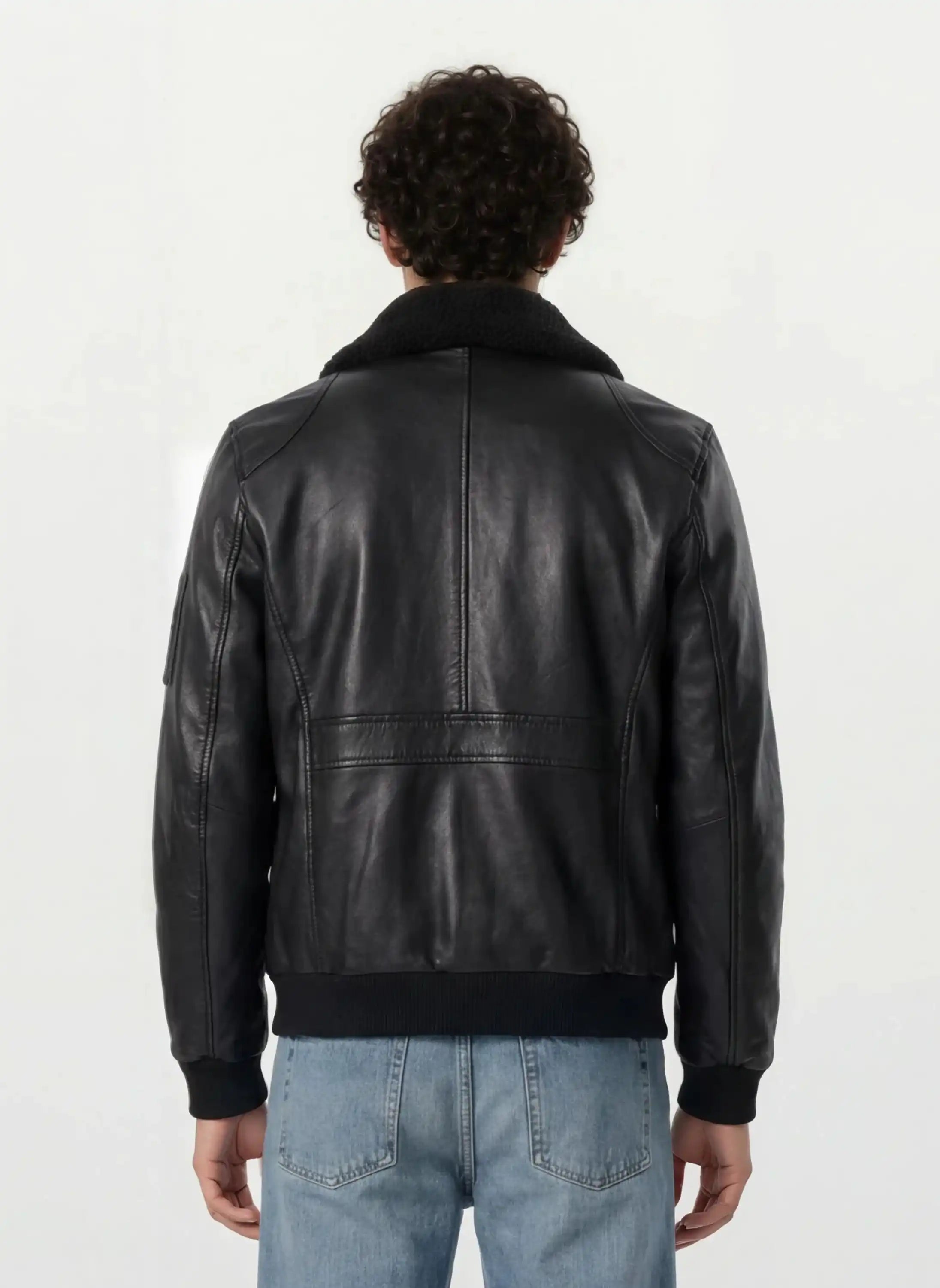 Eine Person mit kurzen lockigen Haaren ist von hinten zu sehen. Sie trägt eine Trueprodigy Moderne Fliegerjacke aus echtem Leder mit abnehmbarem Teddy-Kragen und hellblaue Jeans vor einem schlichten hellen Hintergrund.