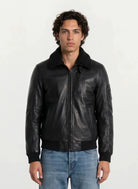 Eine Person mit mittellangem, lockigem Haar trägt eine trueprodigy Moderne Fliegerjacke aus echtem Leder mit abnehmbarem Teddy-Kragen und hellblaue Jeans vor einem schlichten hellen Hintergrund.