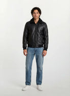 Ein Mann mit kurzen, lockigen dunklen Haaren steht mit dem Gesicht nach vorne vor einem weißen Hintergrund und trägt die trueprodigy Moderne Fliegerjacke aus echtem Leder mit abnehmbarem Teddy-Kragen, hellblaue Jeans und weiße Turnschuhe. Seine Arme ruhen an den Seiten.