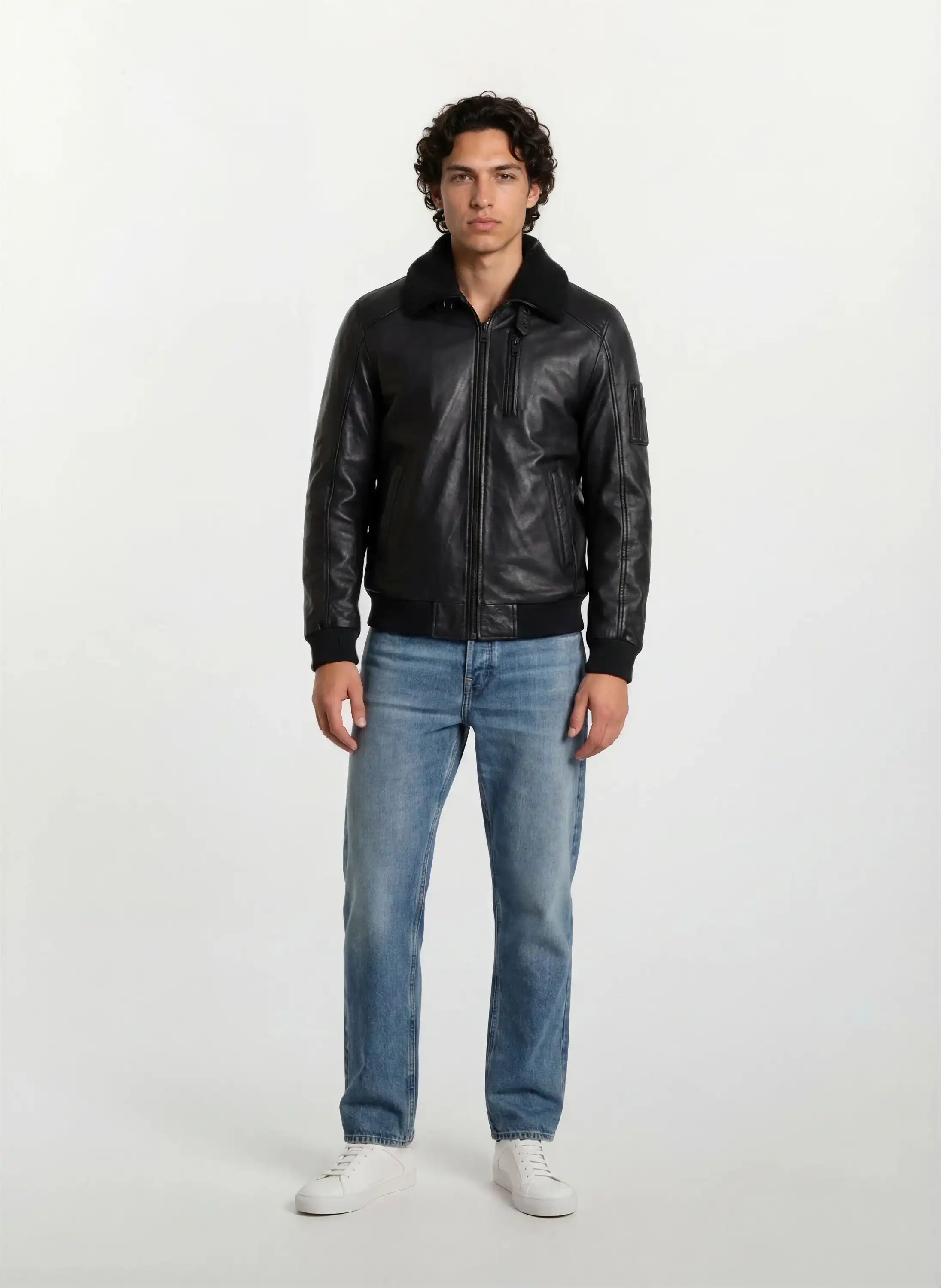 Ein Mann mit kurzen, lockigen dunklen Haaren steht mit dem Gesicht nach vorne vor einem weißen Hintergrund und trägt die trueprodigy Moderne Fliegerjacke aus echtem Leder mit abnehmbarem Teddy-Kragen, hellblaue Jeans und weiße Turnschuhe. Seine Arme ruhen an den Seiten.