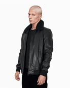 Eine Person mit kurzen, blondierten Haaren trägt eine schwarze Trueprodigy Blaze-Lederjacke mit Lammfellkragen über einem schwarzen Hemd. Sie steht vor einem schlichten weißen Hintergrund und blickt mit neutralem Gesichtsausdruck direkt in die Kamera.