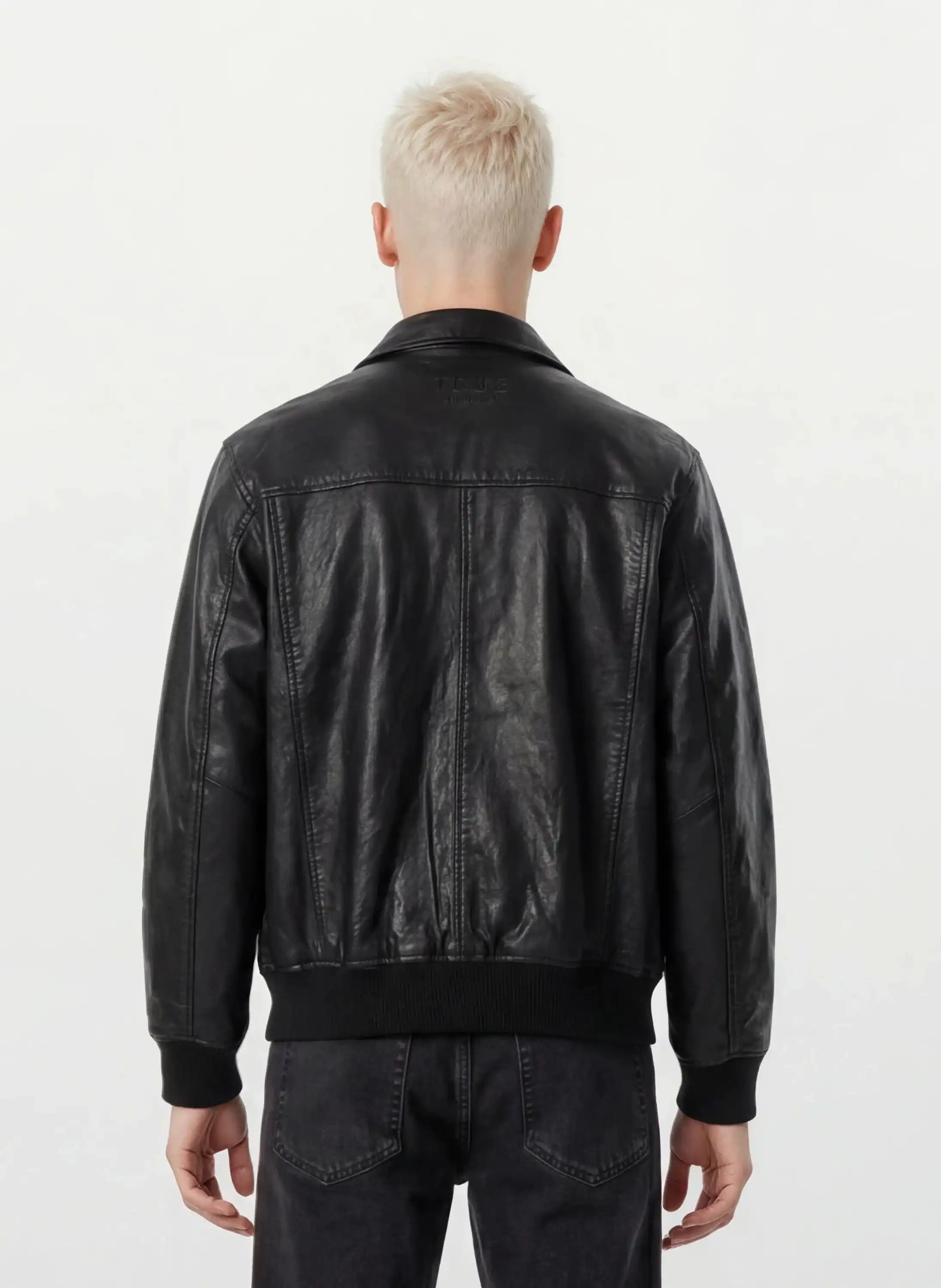 Eine Person mit kurzen, platinblonden Haaren ist von hinten zu sehen und trägt eine trueprodigy Oversize-Lederjacke mit Stehkragen, 3D-Silikonlogo & Retro-Details und dunkle Jeans vor einem schlichten, hellen Hintergrund.