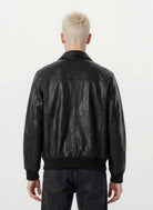 Eine Person mit kurzen, platinblonden Haaren ist von hinten zu sehen und trägt eine trueprodigy Oversize-Lederjacke mit Stehkragen, 3D-Silikonlogo & Retro-Details und dunkle Jeans vor einem schlichten, hellen Hintergrund.