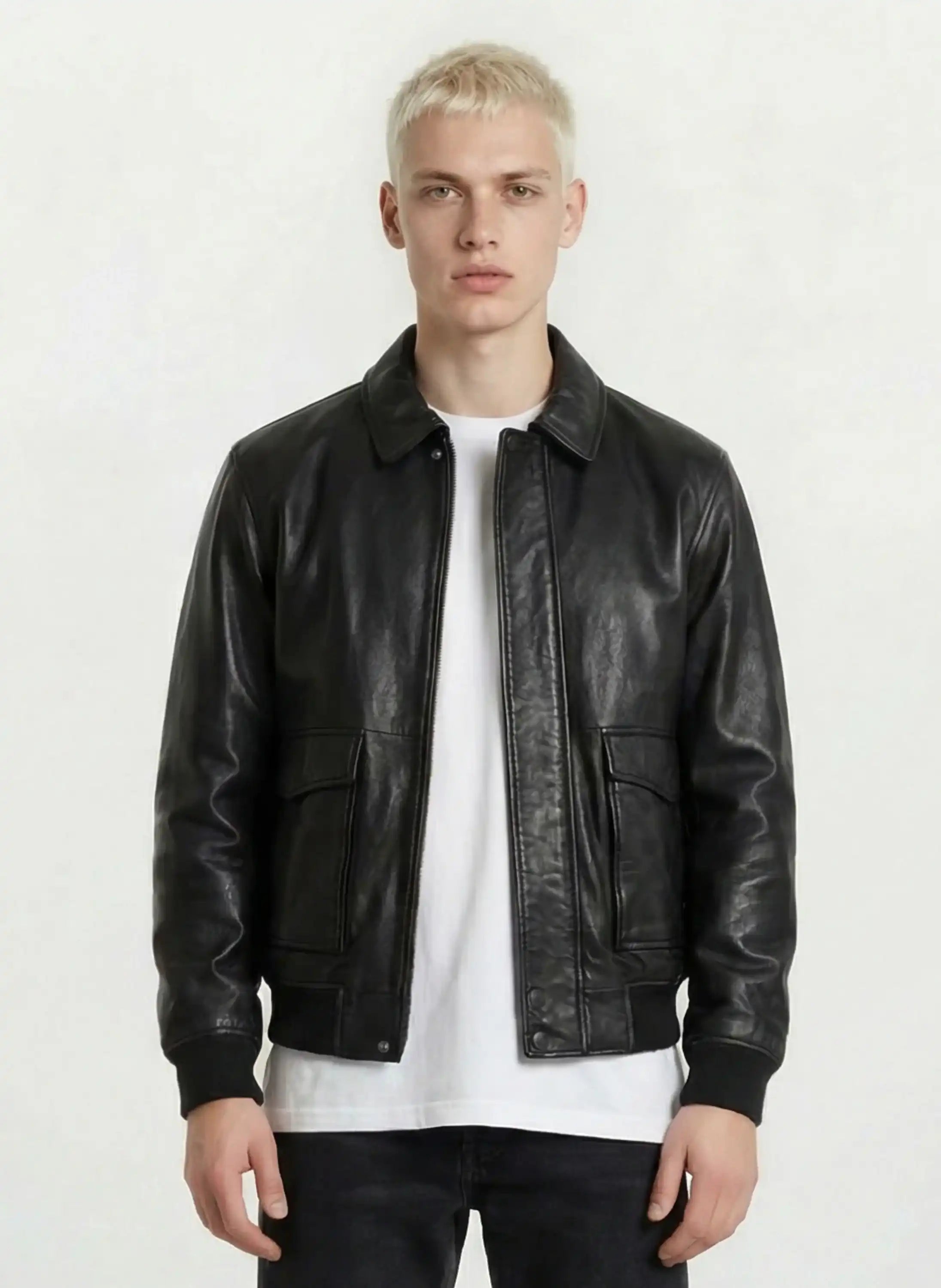 Eine Person mit kurzen blonden Haaren trägt die Oversize-Lederjacke mit Stehkragen, 3D-Silikonlogo & Retro-Details von trueprodigy über einem weißen T-Shirt und einer schwarzen Hose, steht vor einem schlichten hellen Hintergrund und schaut direkt in die Kamera.