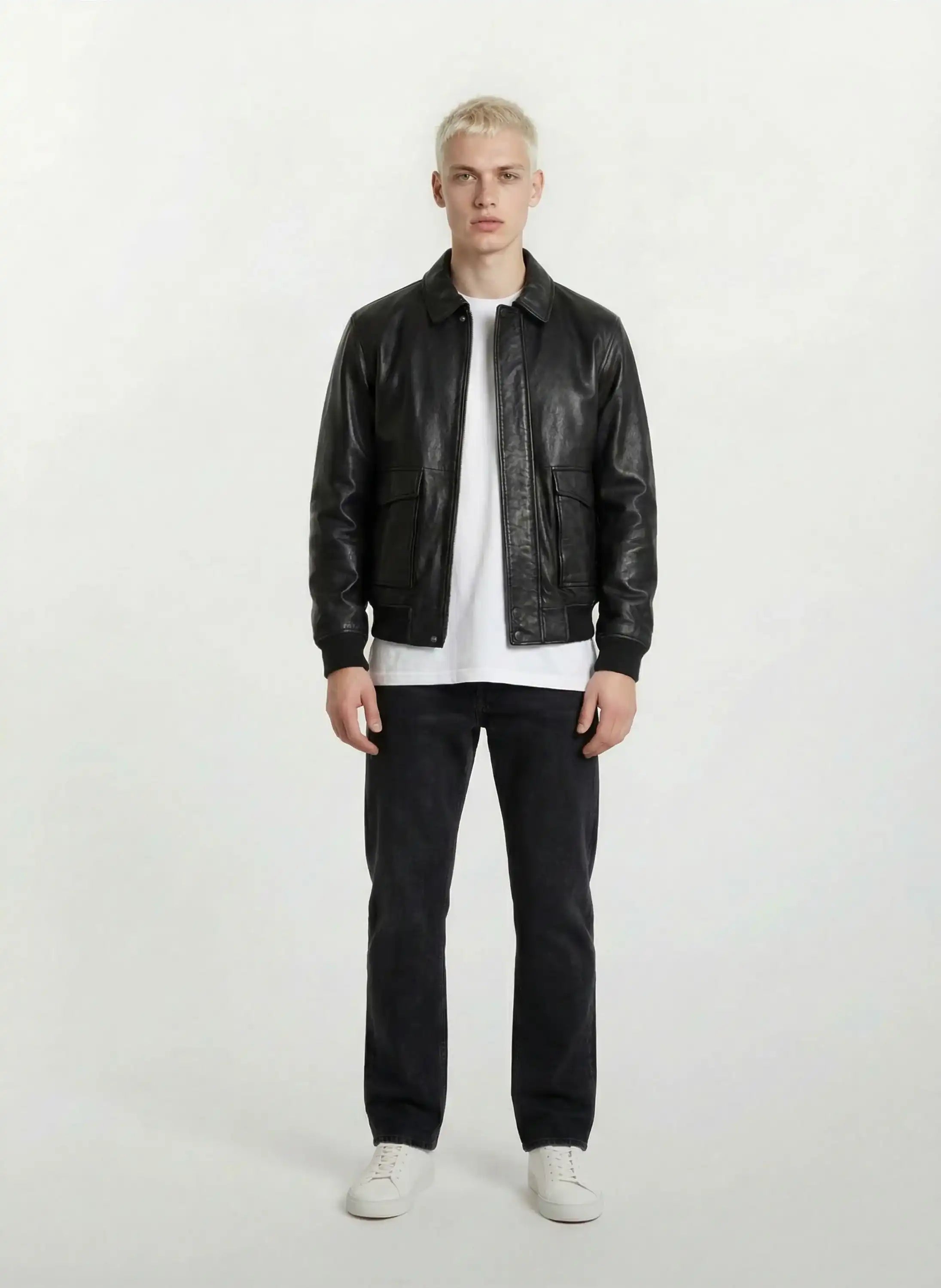Ein junger Mann mit kurzen blonden Haaren schaut nach vorne in einer Trueprodigy Oversize-Lederjacke mit Stehkragen, 3D-Silikonlogo & Retro-Details, gestylt mit einem weißen T-Shirt, schwarzen Jeans und weißen Turnschuhen vor einem schlichten hellen Hintergrund.