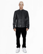 Eine Person mit kurzen, hellen Haaren steht vor einem weißen Hintergrund. Sie trägt Dash – eine elegante schwarze Lederjacke von trueprodigy mit Stehkragen. Dazu eine schwarze Hose und weiße Turnschuhe. Die Hände hängen an den Seiten und der Blick ist direkt in die Kamera gerichtet.