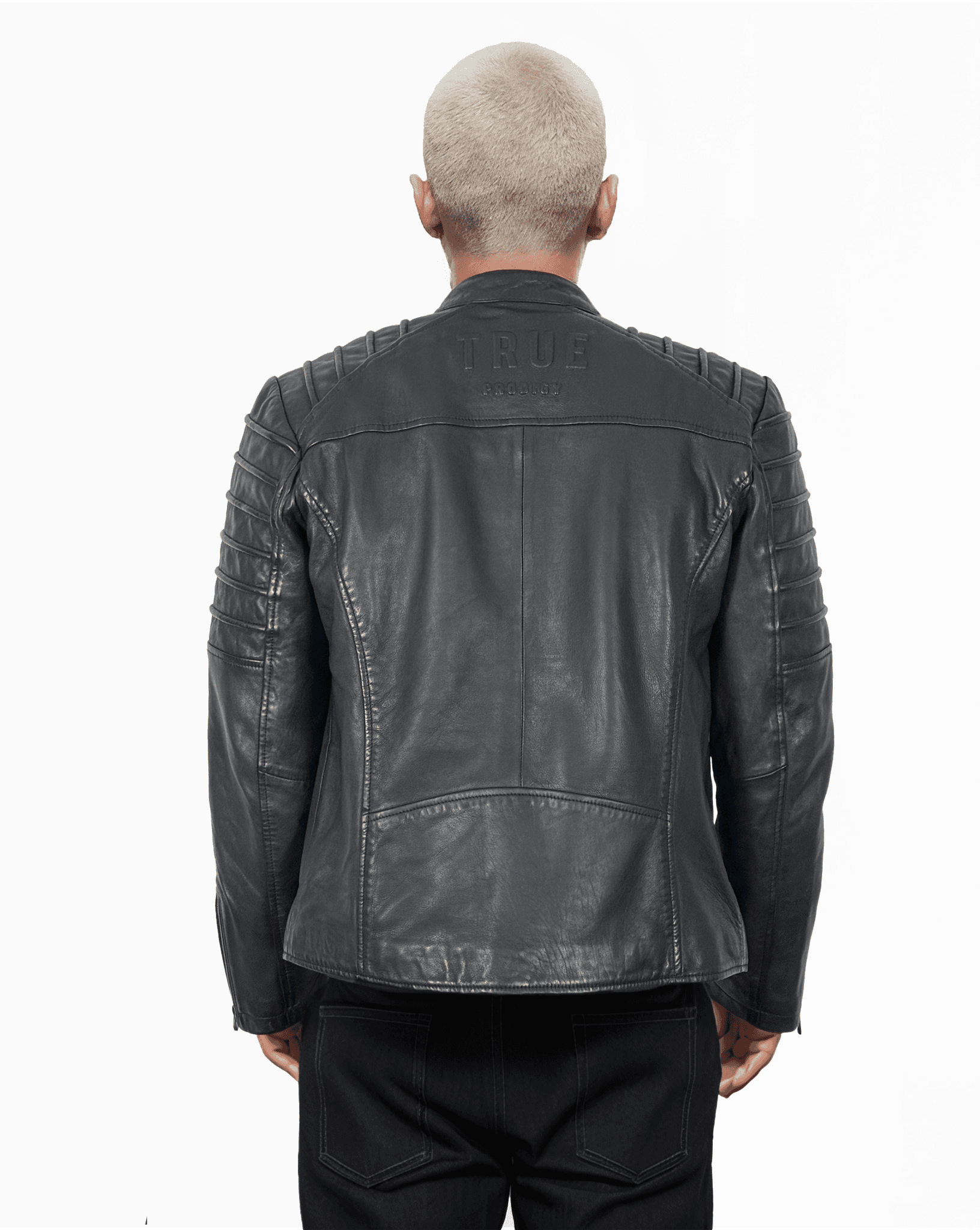 Eine Person mit kurzen, hellen Haaren ist von hinten zu sehen. Sie trägt eine schwarze Trueprodigy Dash-Jacke mit gesteppten Schulterdetails und dem Schriftzug „TRUE“ oben. Die Jacke hat einen eleganten Stehkragen, der perfekt zur dunklen Hose passt.