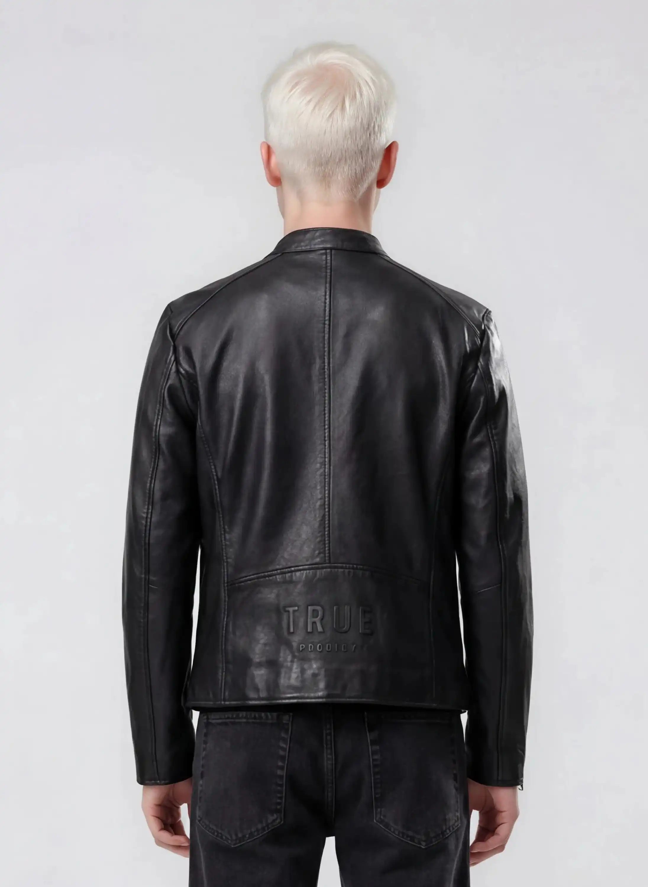 Eine Person mit kurzem platinblondem Haar, von hinten gesehen in schwarzer Jeans und einer trueprodigy Slim-Fit-Lederjacke mit sichtbarem 3D-Rückenlogo aus Schafsleder. Das Wort TRUE ist auf dem unteren Rücken der Jacke geprägt. Der Hintergrund ist hellgrau.