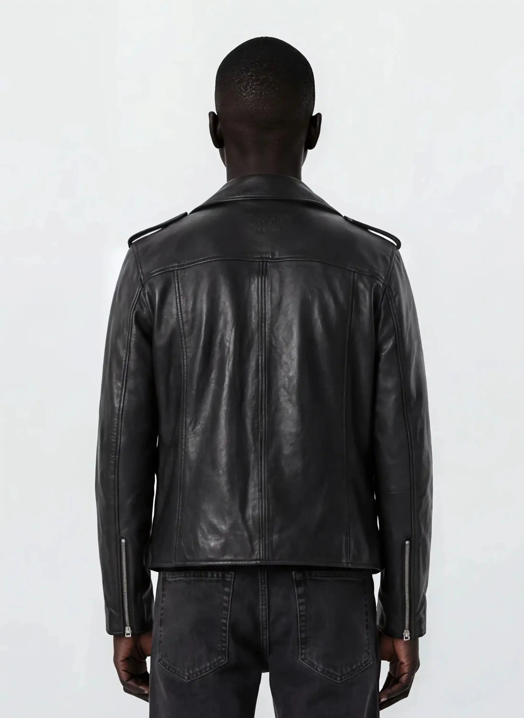 Eine Person mit kurzen Haaren trägt eine trueprodigy Leder-Bikerjacke mit asymmetrischem Zipper & 3D-Logo unter dem Kragen und schwarze Jeans, steht mit dem Rücken zur Kamera und präsentiert einen coolen Biker-Look vor einem schlichten hellen Hintergrund.