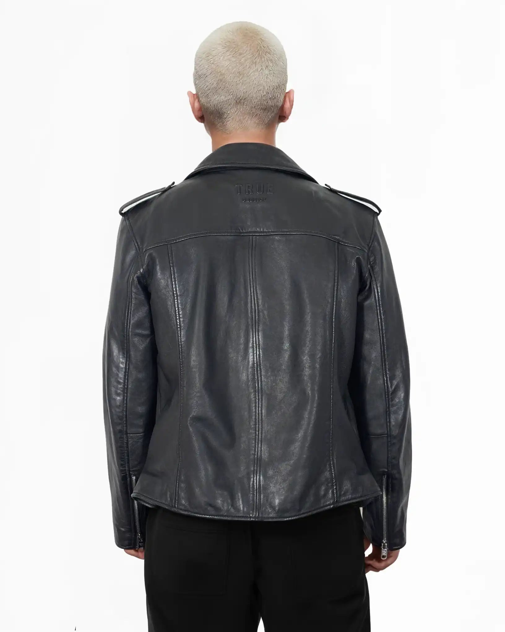 Eine Person mit kurzen, gebleichten Haaren ist von hinten zu sehen. Sie trägt die trueprodigy Easton Leder-Bikerjacke mit asymmetrischem Zipper & 3D-Logo unter dem Kragen und schwarzer Hose und setzt damit einen coolen Biker-Look vor einem schlichten weißen Hintergrund um.