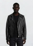 Eine Person blickt in die Kamera und trägt eine trueprodigy Leder-Bikerjacke mit asymmetrischem Zipper & 3D-Logo unter dem Kragen über einem schwarzen Hemd und einer dunklen Jeans vor einem schlichten hellgrauen Hintergrund.