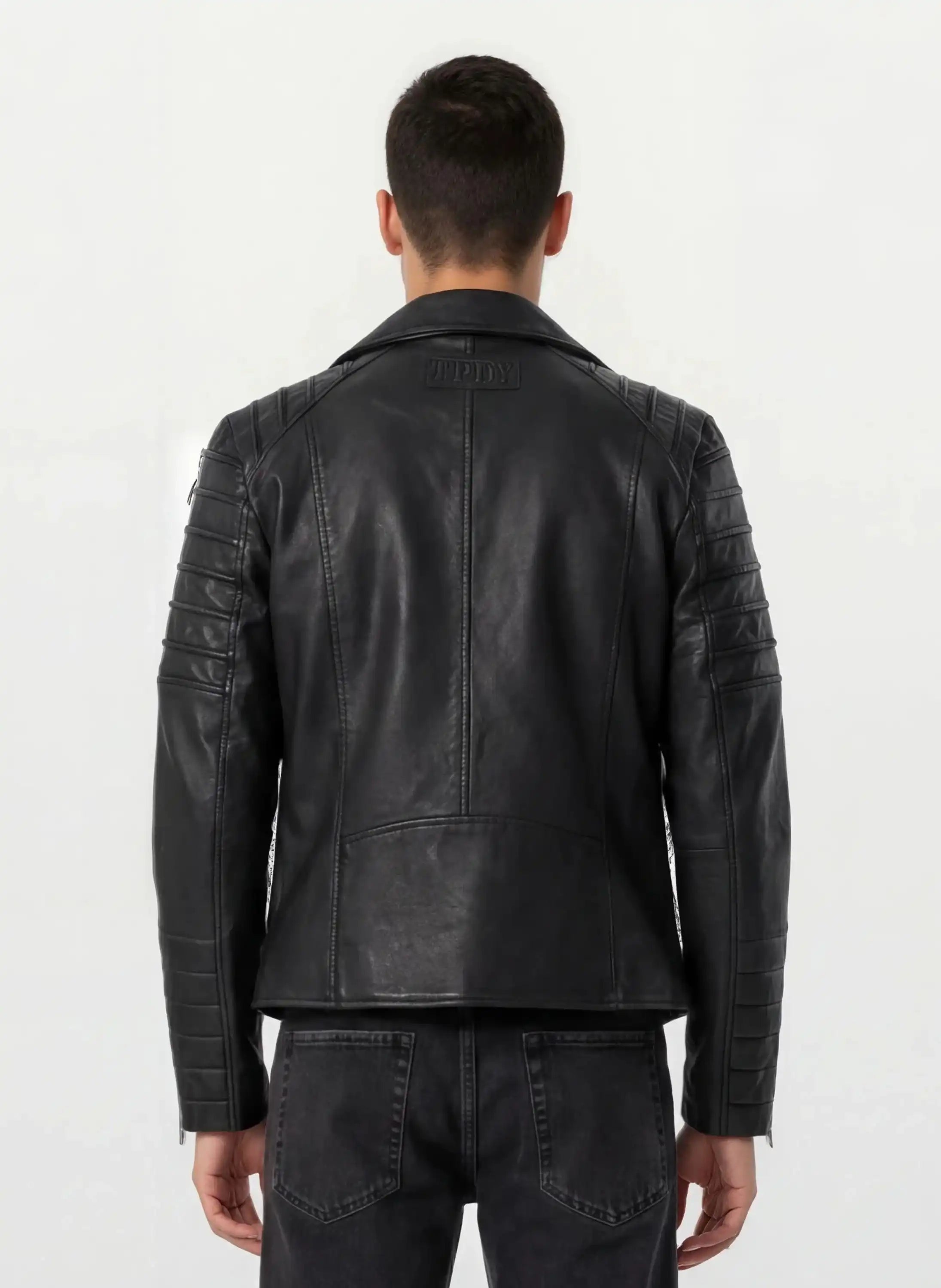 Ein Mann ist von hinten zu sehen, der eine Trueprodigy Lederjacke mit Paspelierung und diversen Reißverschlüssen trägt, gepaart mit einer dunklen Jeans vor einem schlichten hellen Hintergrund.