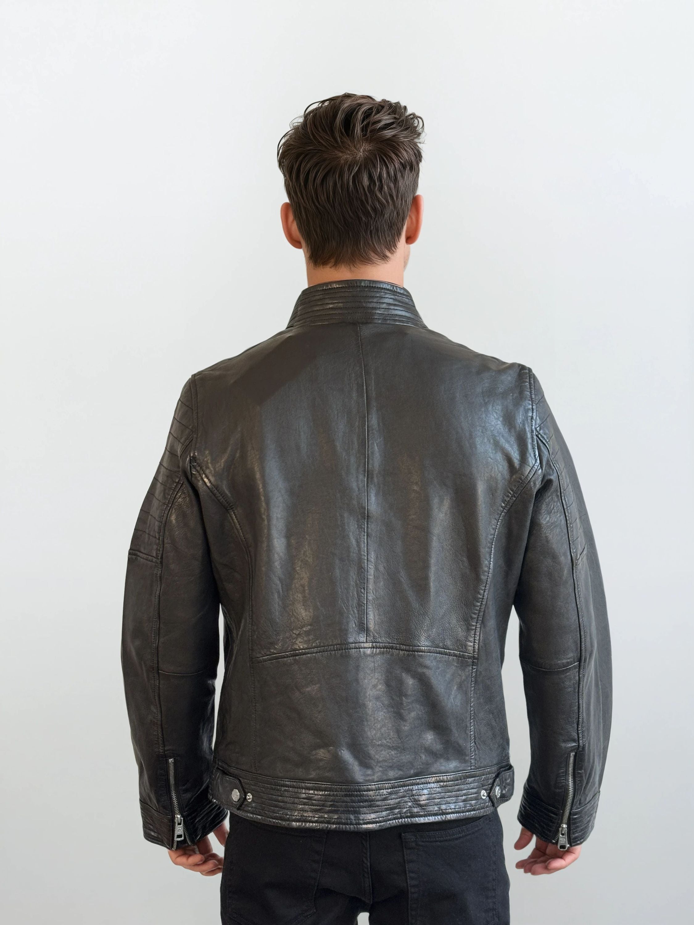 Eine Person mit kurzen braunen Haaren ist von hinten zu sehen, die eine trueprodigy Herren Lederjacke aus Schafsleder in Schwarz mit klassischem Blouson-Schnitt und eine schwarze Hose vor einem schlichten hellen Hintergrund trägt.