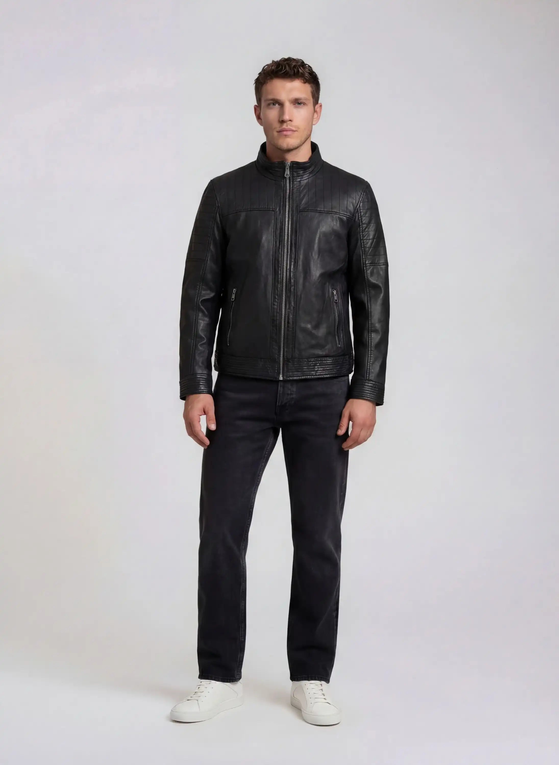 Ein Mann trägt die schwarze trueprodigy Herren Lederjacke aus Schafsleder im klassischen Blouson-Schnitt, kombiniert mit dunkler Jeans und weißen Sneakers. Seine Arme hängen locker an den Seiten, während er vor einem hellen Hintergrund steht.