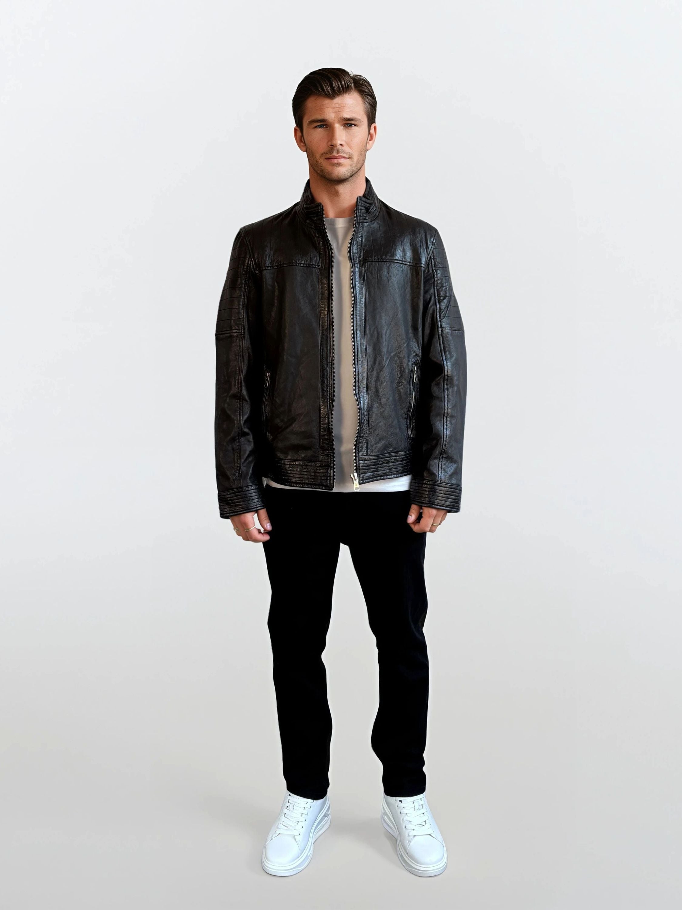 Ein Mann trägt die trueprodigy Herren Lederjacke aus Schafsleder in Schwarz mit klassischem Blouson-Schnitt über einem beigen Shirt, kombiniert mit schwarzen Hosen und weißen Sneakern vor hellem Hintergrund.