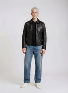 Eine Person mit kurzen blonden Haaren steht mit dem Gesicht nach vorne und trägt eine trueprodigy Herren Lederjacke aus Schafsleder in Schwarz mit Hemd-Design, gepaart mit einem schwarzen Hemd, blauen Jeans und weißen Turnschuhen vor einem schlichten hellen Hintergrund.