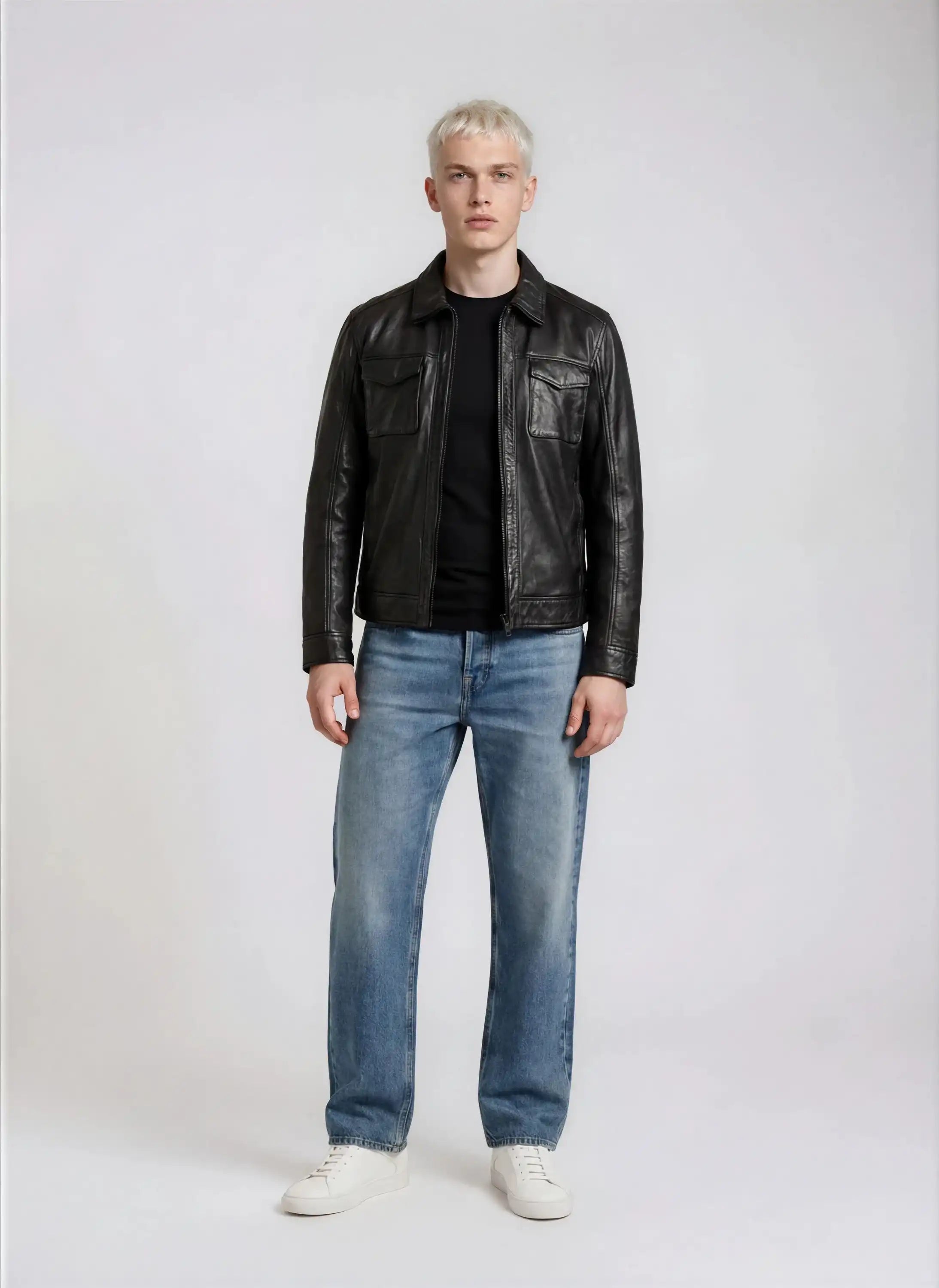 Eine Person mit kurzen blonden Haaren steht mit dem Gesicht nach vorne und trägt eine trueprodigy Herren Lederjacke aus Schafsleder in Schwarz mit Hemd-Design, gepaart mit einem schwarzen Hemd, blauen Jeans und weißen Turnschuhen vor einem schlichten hellen Hintergrund.