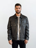 Ein Mann mit kurzen dunklen Haaren und Bart steht vor einem hellen Hintergrund, trägt die trueprodigy Herren Lederjacke aus Schafsleder in Schwarz mit Hemd-Design über einem weißen Hemd und einer schwarzen Hose und schaut neutral in die Kamera.