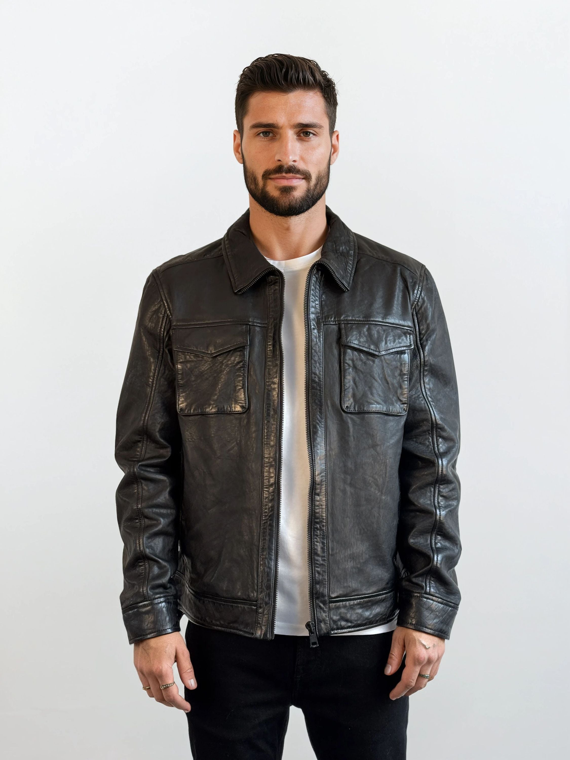Ein Mann mit kurzen dunklen Haaren und Bart steht vor einem hellen Hintergrund, trägt die trueprodigy Herren Lederjacke aus Schafsleder in Schwarz mit Hemd-Design über einem weißen Hemd und einer schwarzen Hose und schaut neutral in die Kamera.