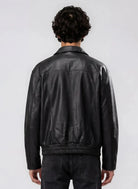 Gezeigt wird eine Person mit lockigem dunklem Haar von hinten, die die trueprodigy Herren Lederjacke aus Schafsleder in Schwarz mit klassischem Blouson-Schnitt und schwarzer Jeans trägt und vor einem schlichten hellgrauen Hintergrund steht.