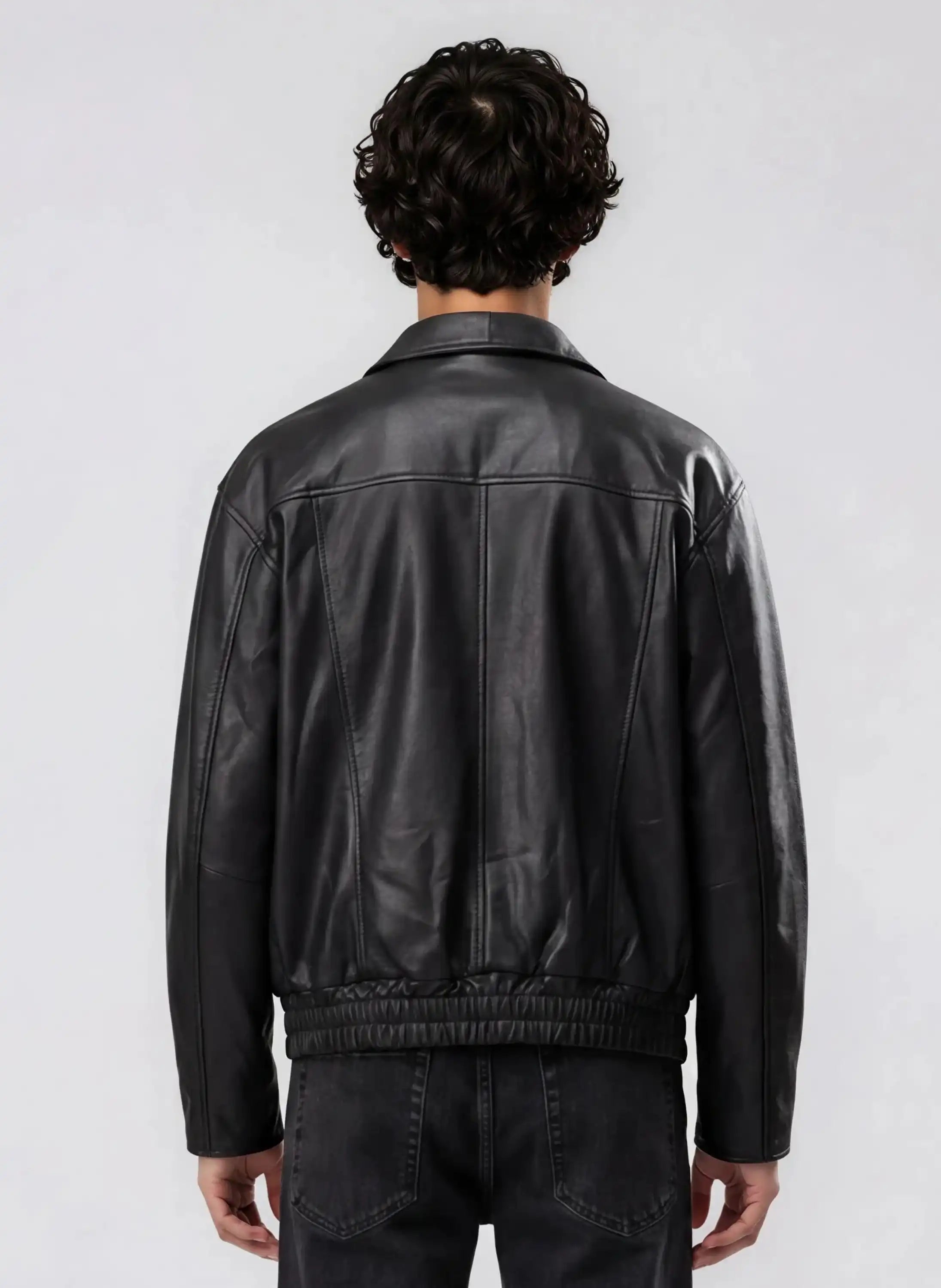 Gezeigt wird eine Person mit lockigem dunklem Haar von hinten, die die trueprodigy Herren Lederjacke aus Schafsleder in Schwarz mit klassischem Blouson-Schnitt und schwarzer Jeans trägt und vor einem schlichten hellgrauen Hintergrund steht.