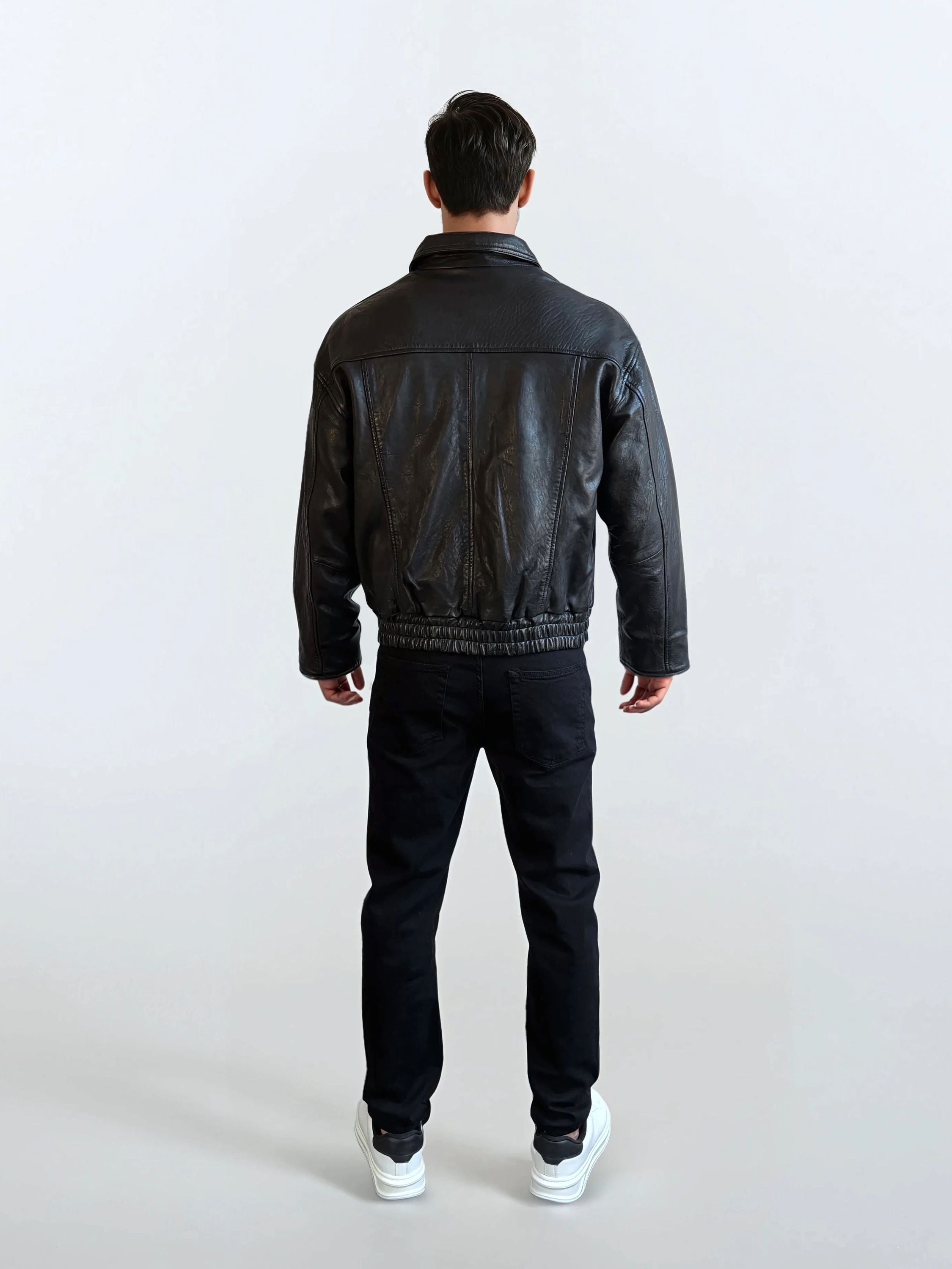 Eine Person blickt von der Kamera weg und trägt die trueprodigy Herren Lederjacke aus Schafsleder in Schwarz mit klassischem Blouson-Schnitt, eine schwarze Hose und weiße Turnschuhe vor einem schlichten hellen Hintergrund.