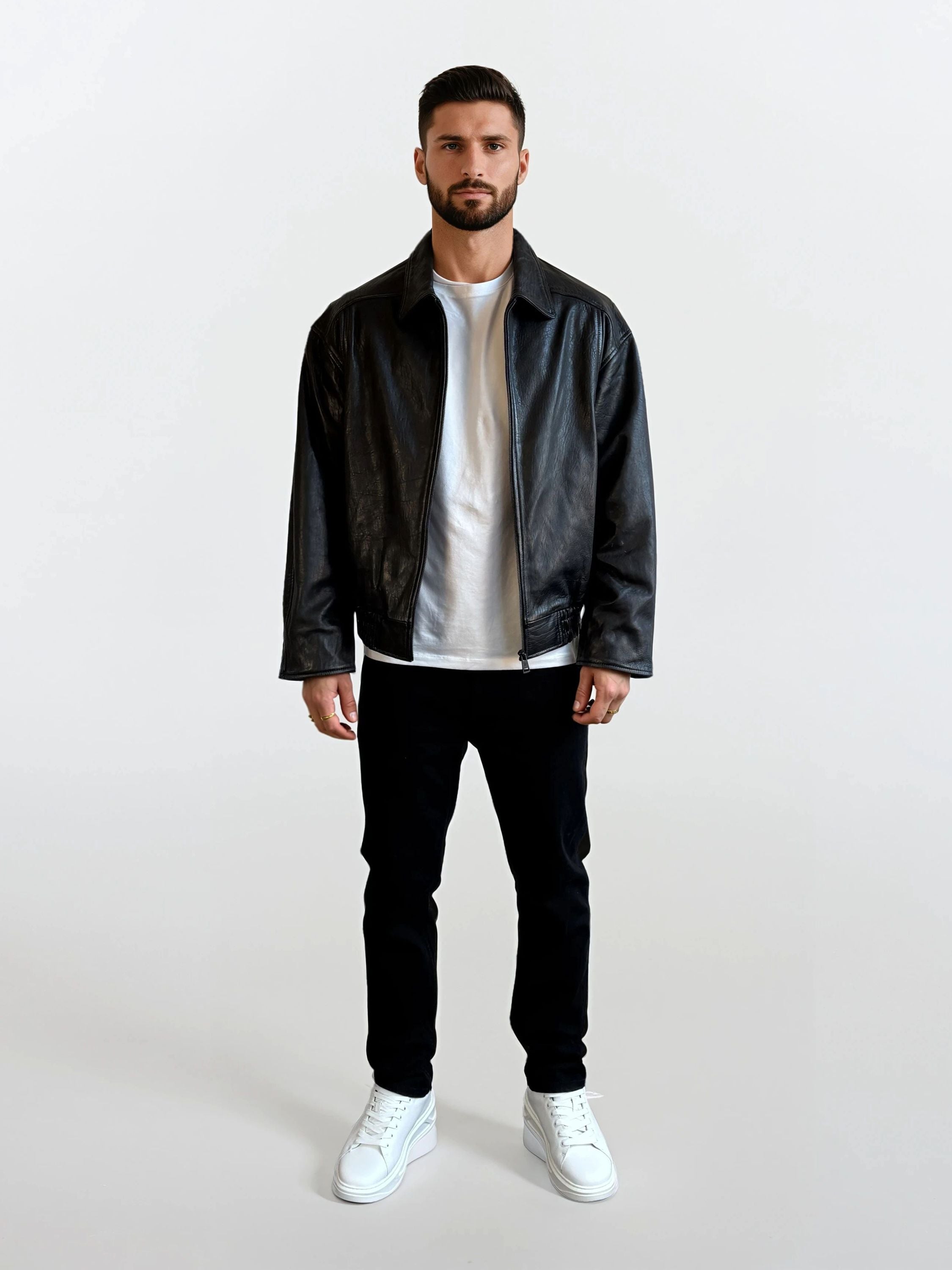 Ein Mann mit kurzen dunklen Haaren und einem Bart, der eine trueprodigy Herren Lederjacke aus Schafsleder in Schwarz mit klassischem Blouson-Schnitt, ein weißes T-Shirt, eine schwarze Hose und weiße Turnschuhe trägt, steht vor einem schlichten weißen Hintergrund.