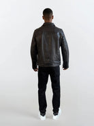 Eine Person trägt eine schwarze Herren Lederjacke aus Schafsleder mit Ärmel-Reißverschlüssen von trueprodigy, dazu schwarze Hosen und weiße Sneaker vor einem schlichten, hellen Hintergrund.