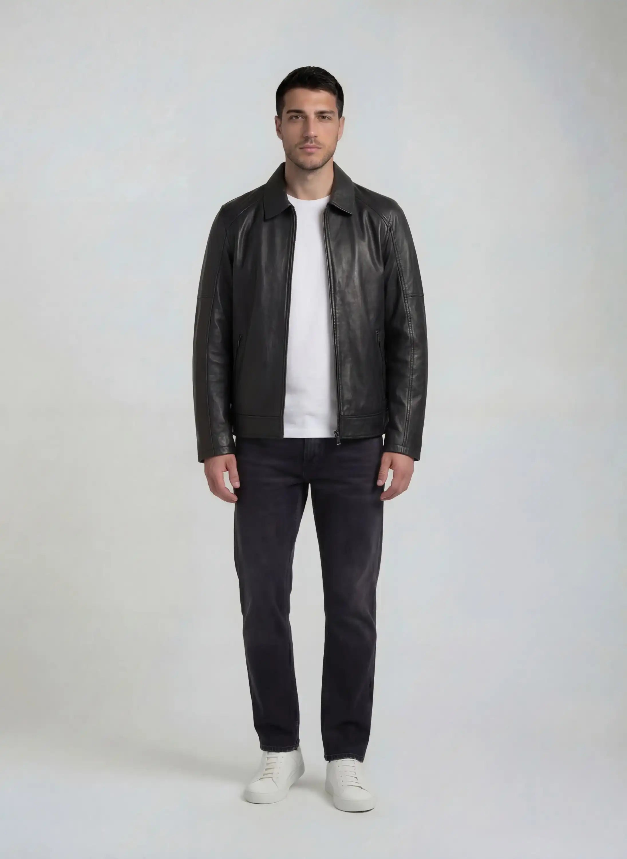 Ein Mann trägt eine schwarze Herren Lederjacke aus Schafsleder mit Ärmel-Reißverschlüssen von trueprodigy über einem weißen T-Shirt, dunkle Jeans und weiße Sneaker vor einem schlichten hellgrauen Hintergrund.