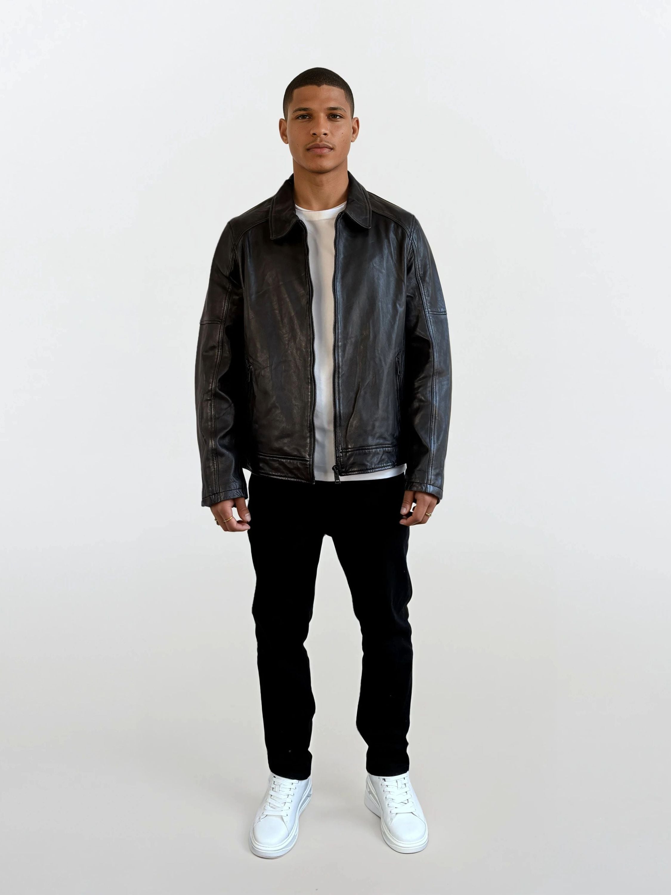 Eine Person steht vor weißem Hintergrund und trägt eine schwarze Herren Lederjacke aus Schafsleder mit Ärmel-Reißverschlüssen von trueprodigy, dazu ein weißes T-Shirt, schwarze Hosen und weiße Sneakers.