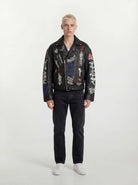 Eine Person mit kurzen blonden Haaren schaut nach vorne und trägt eine trueprodigy Herren Lederjacke aus Schafsleder in Schwarz mit Graffiti-Prints, gepaart mit schwarzen Jeans und weißen Turnschuhen vor einem schlichten hellen Hintergrund.