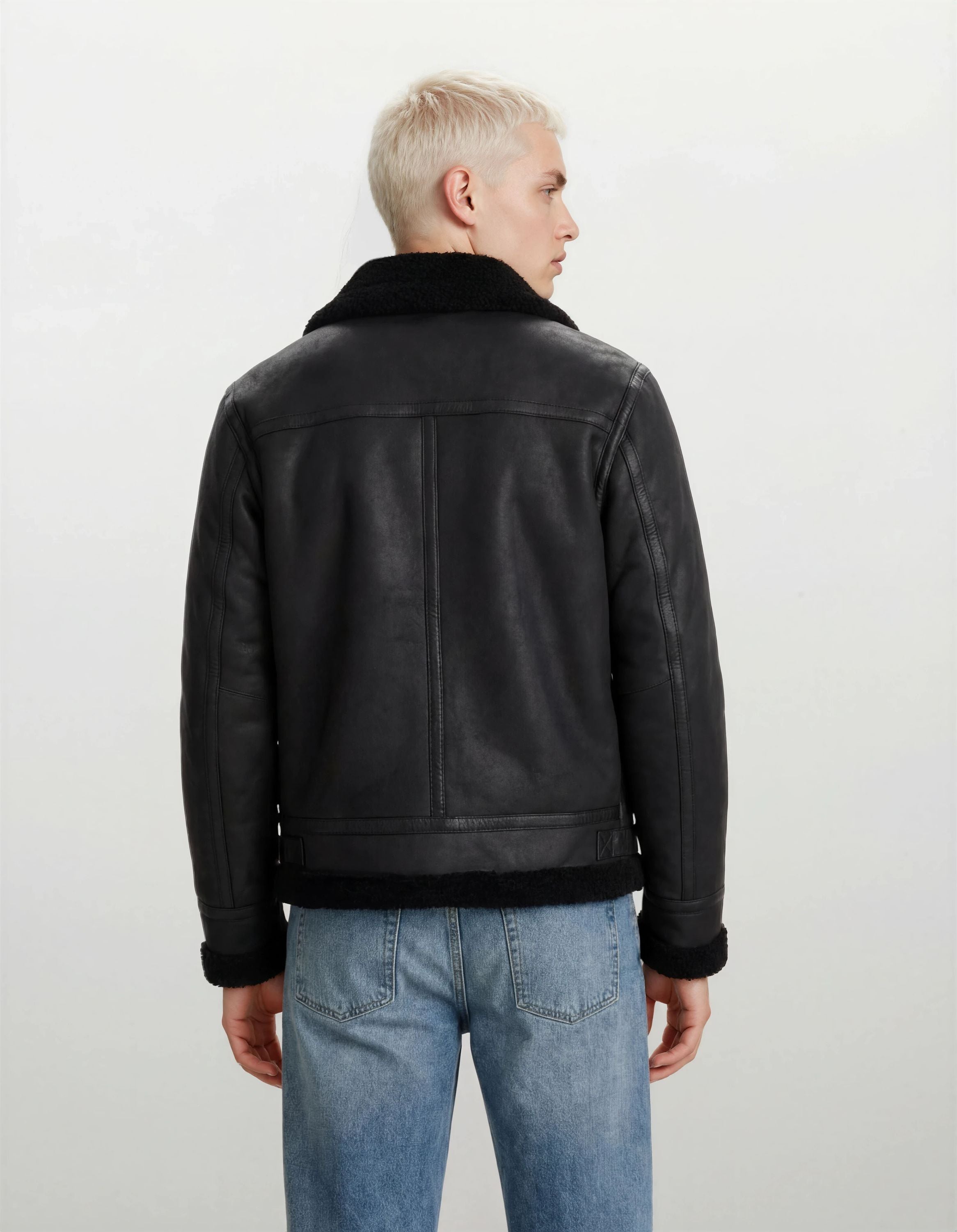 Man sieht eine Person mit kurzen, hellblonden Haaren von hinten, die eine trueprodigy Herren Lederjacke aus Schafsleder in Schwarz im Aviator-Stil mit Fellbesatz und hellblaue Jeans vor einem schlichten hellen Hintergrund trägt.