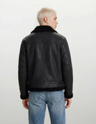 Man sieht eine Person mit kurzen, hellblonden Haaren von hinten, die eine trueprodigy Herren Lederjacke aus Schafsleder in Schwarz im Aviator-Stil mit Fellbesatz und hellblaue Jeans vor einem schlichten hellen Hintergrund trägt.