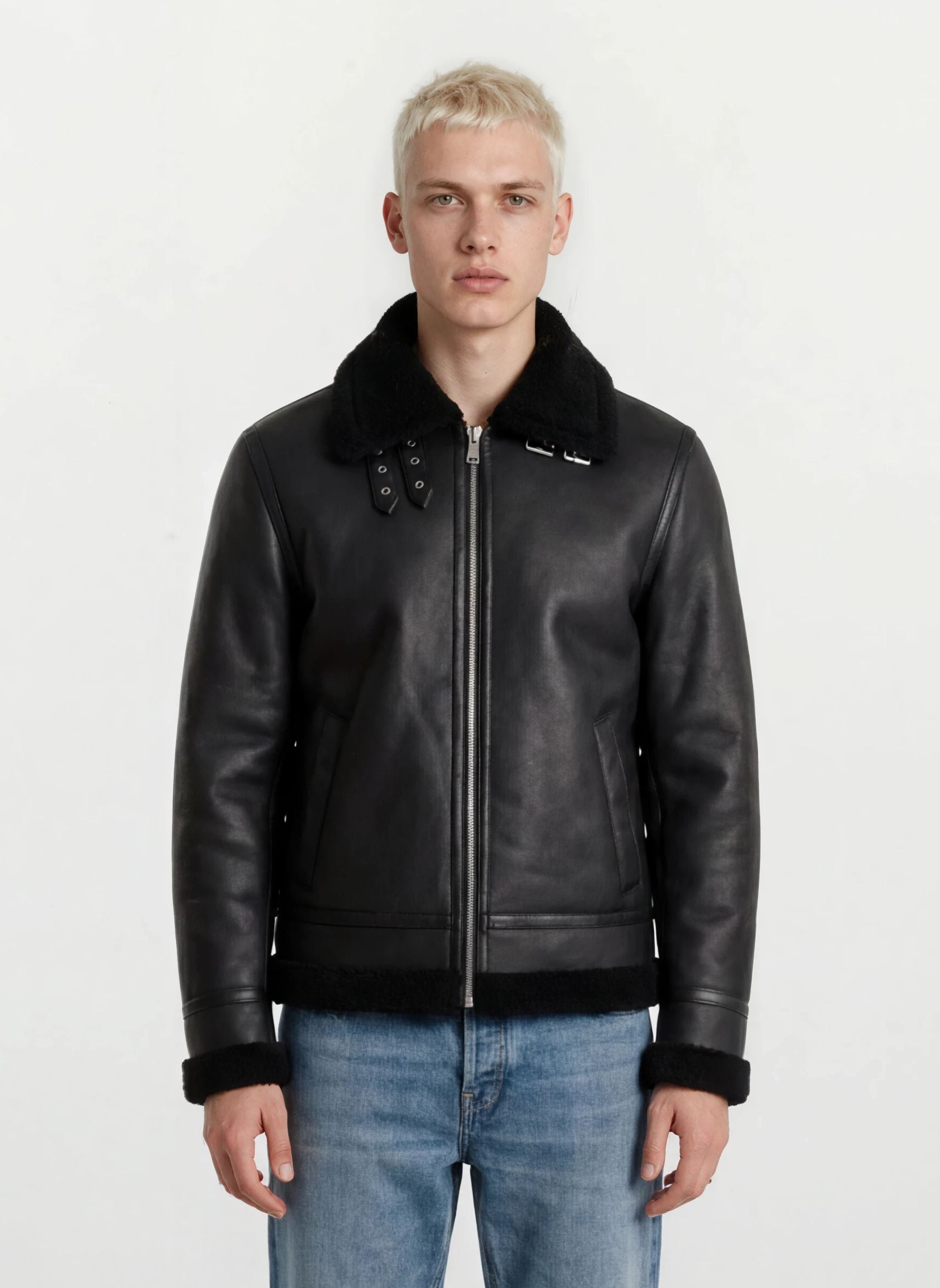 Eine Person mit kurzen blonden Haaren trägt die trueprodigy Herren Lederjacke aus Schafsleder in Schwarz im Aviator-Stil mit Fellbesatz und blaue Jeans, stehend vor einem schlichten weißen Hintergrund.