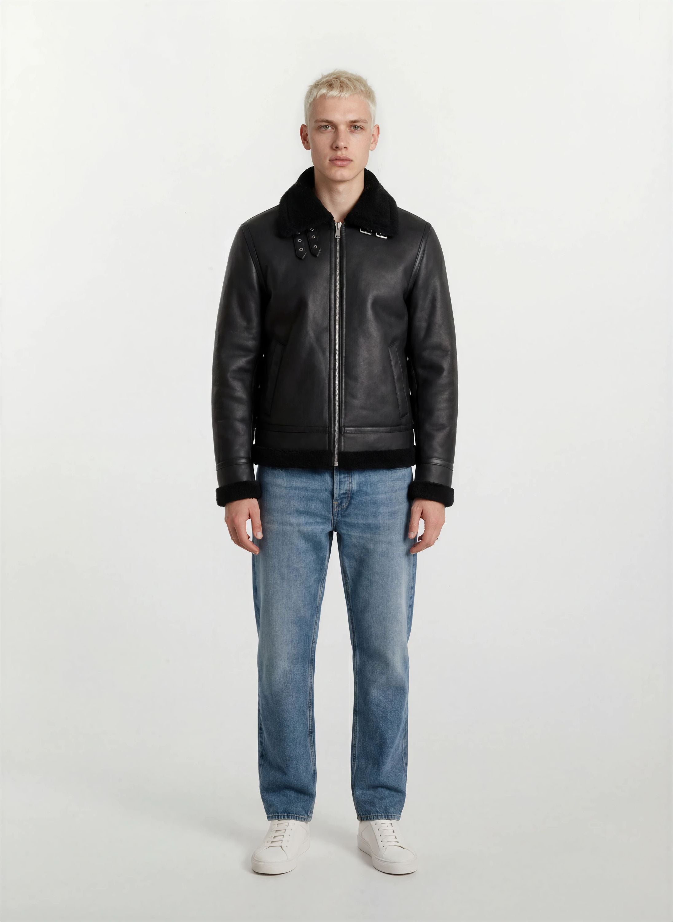 Eine Person mit kurzen blonden Haaren schaut nach vorne und trägt die trueprodigy Herren Lederjacke aus Schafsleder in Schwarz im Aviator-Stil mit Fellbesatz, gepaart mit hellblauen Jeans und weißen Turnschuhen vor einem schlichten hellen Hintergrund.